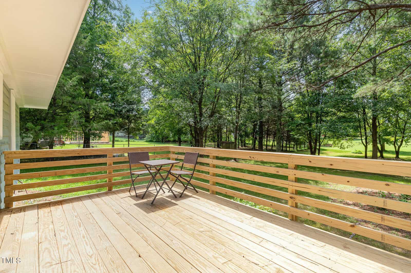 A wooden deck with a small table and two chairs overlooks a grassy yard with tall green trees. Sunlight filters through the trees, casting shadows on the deck.
