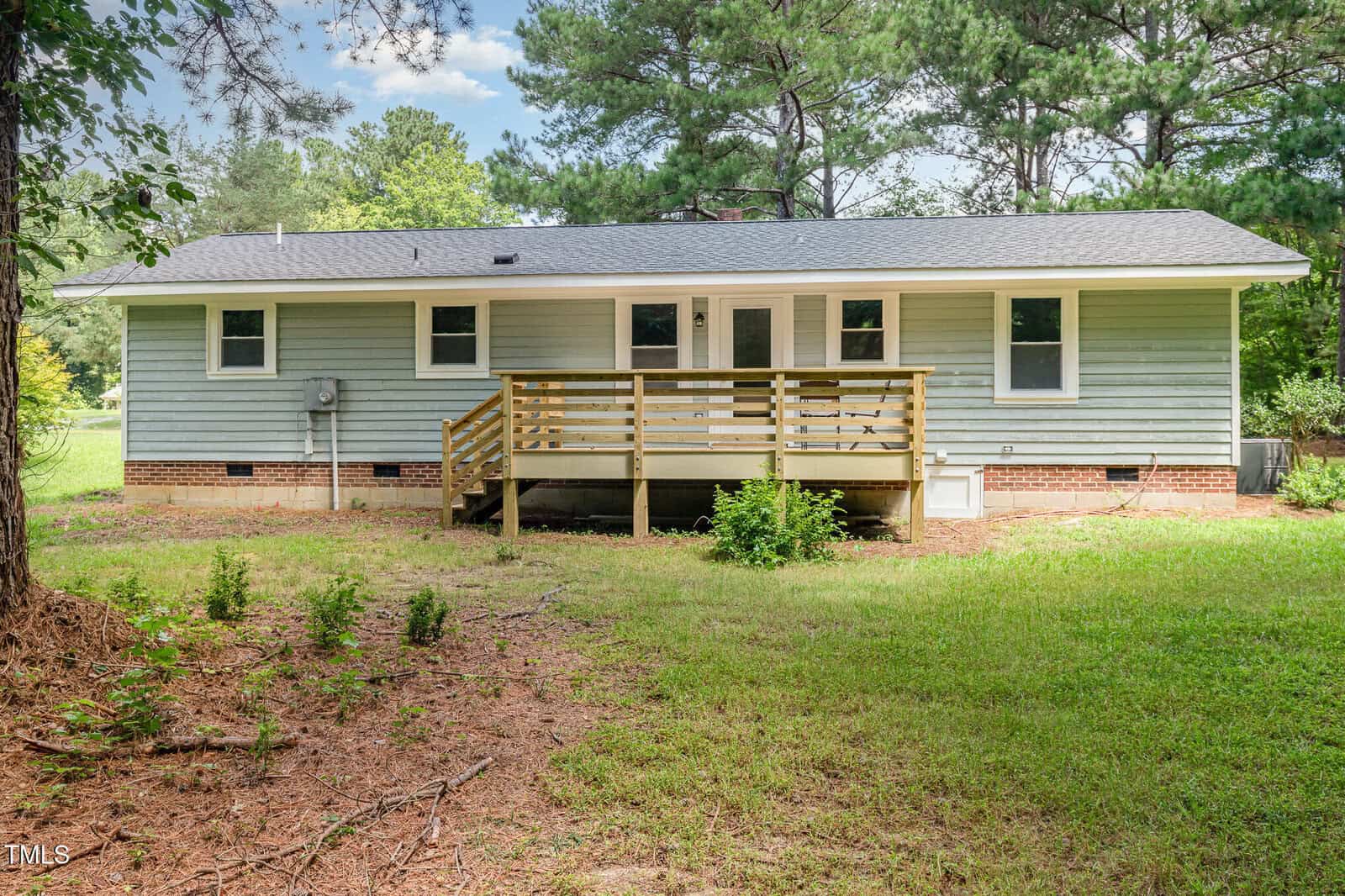 Single-story house with light blue siding, a gray shingled roof, white trim, and a wooden deck with stairs leading to a grassy backyard surrounded by trees.