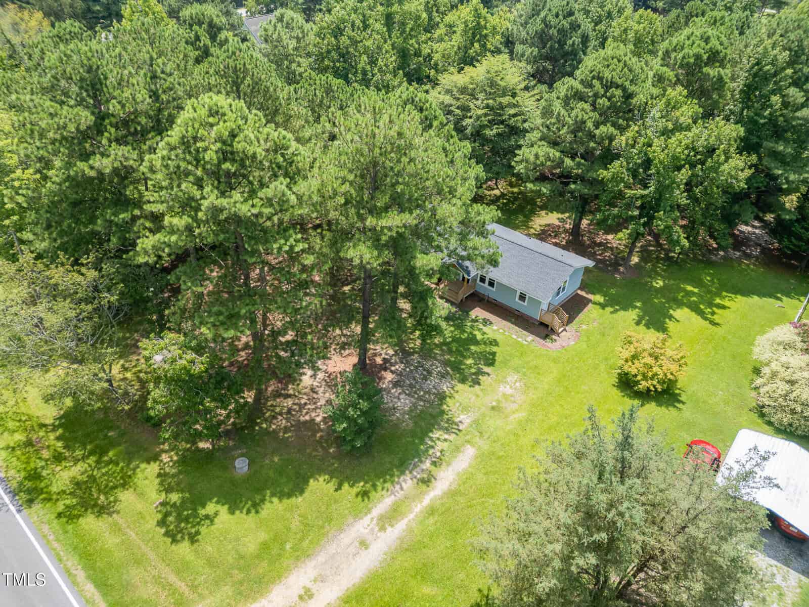 Aerial view of a small house surrounded by tall trees and green grass, located near a road with a driveway and partial view of a red vehicle and a white structure in the lower right corner.