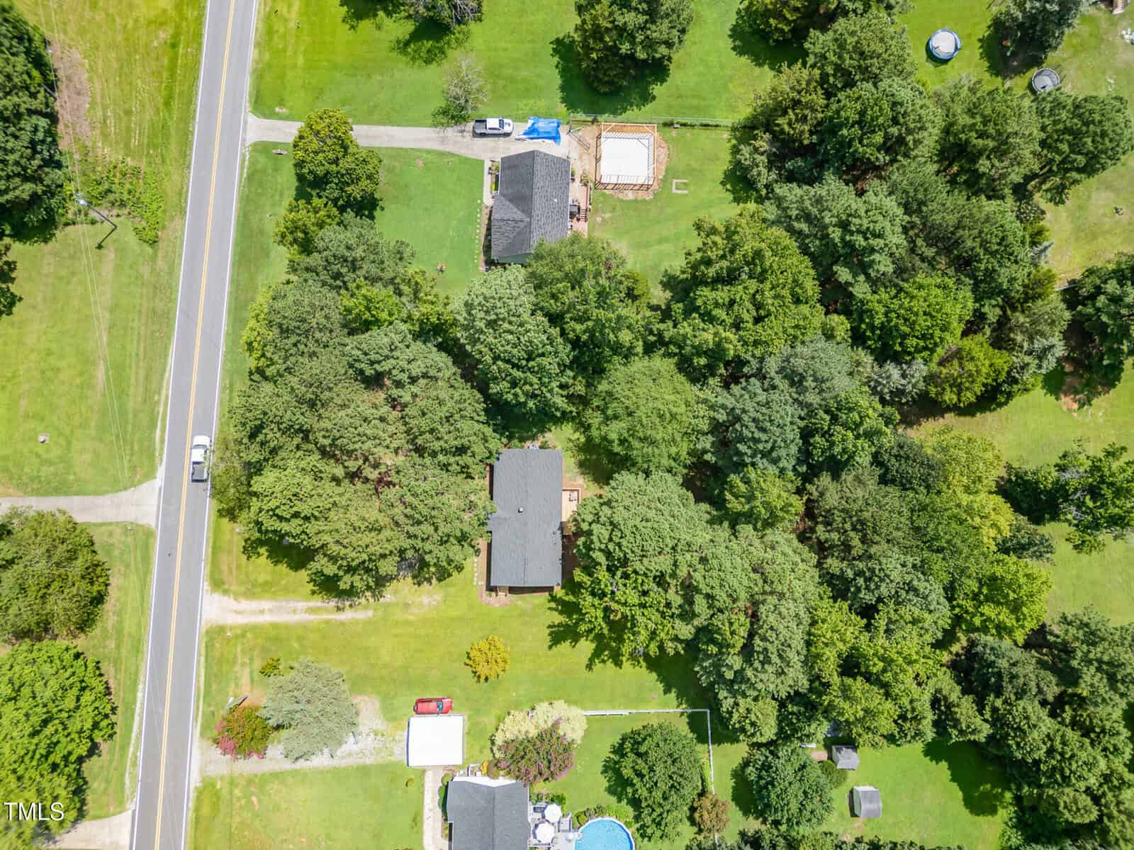Aerial view of a suburban neighborhood with two houses surrounded by trees, a road on the left, and a swimming pool in a backyard near the bottom of the image. Green lawns and scattered vehicles are also visible.