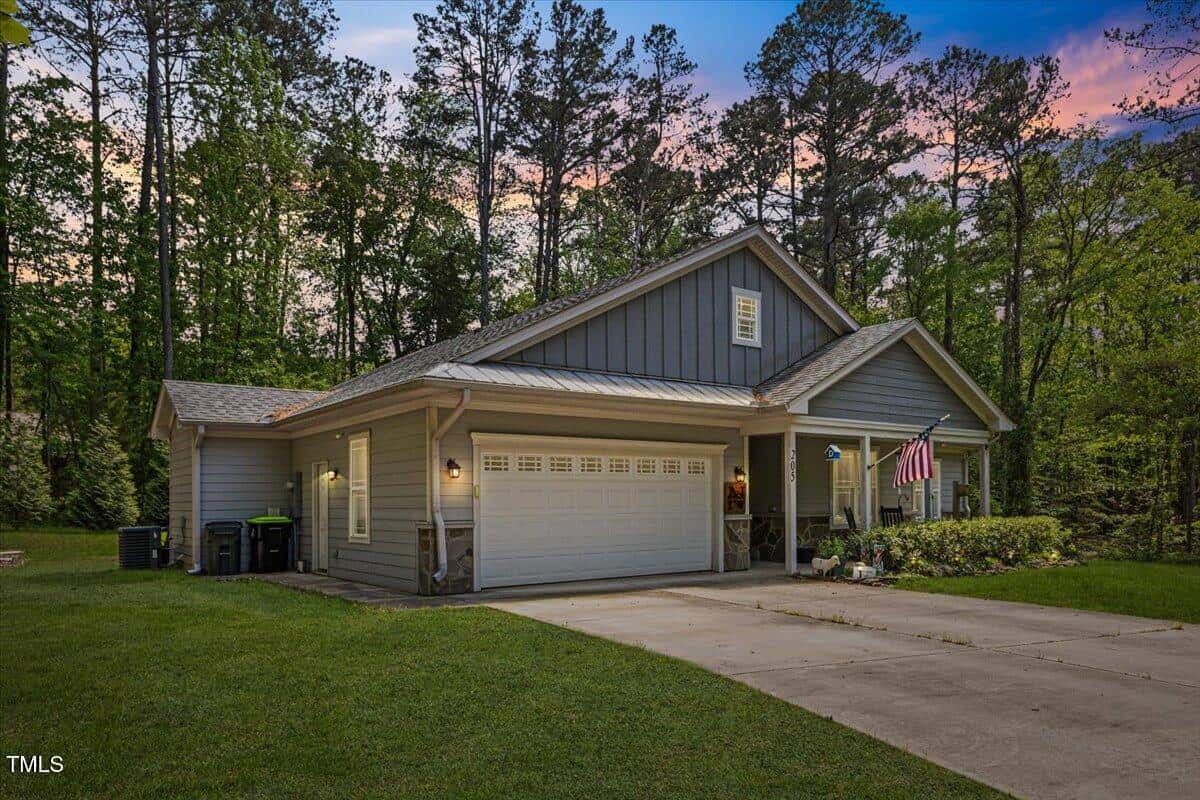 A single-story gray house with a double garage, American and another flag hanging by the entrance, set among tall trees and greenery at dusk. A concrete driveway leads up to the garage.