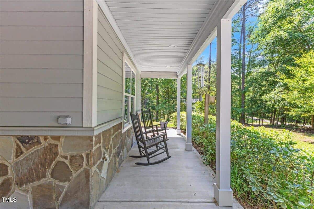 A covered porch with two black rocking chairs on a concrete floor. The house has light gray siding with a stone base and white columns. Green shrubs line the porch, and trees are visible in the background.