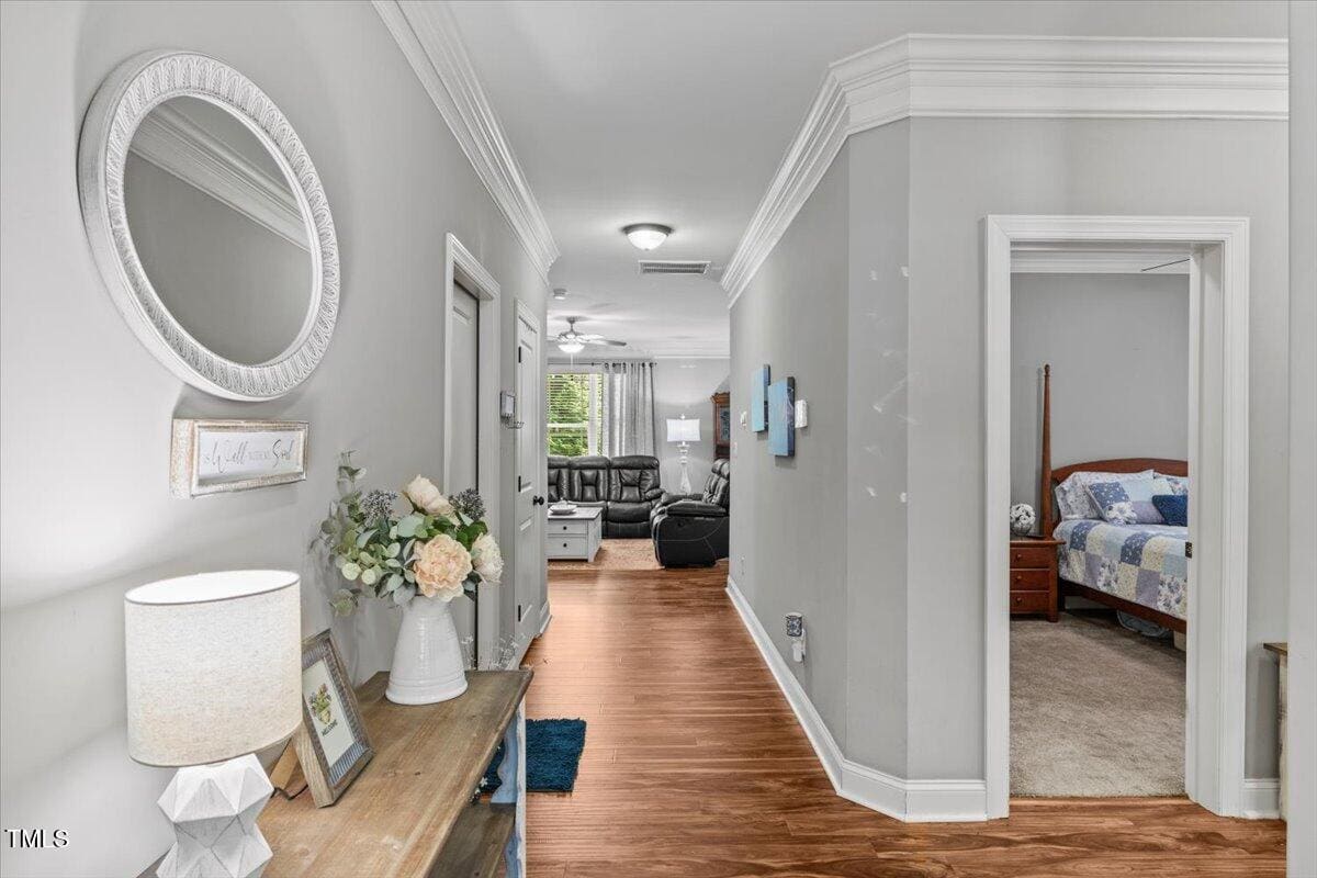 A hallway with wood flooring features a console table holding a lamp and flower arrangement, a round mirror above, and doors leading to a bedroom with a patterned bedspread and a living room with sofas in the background.