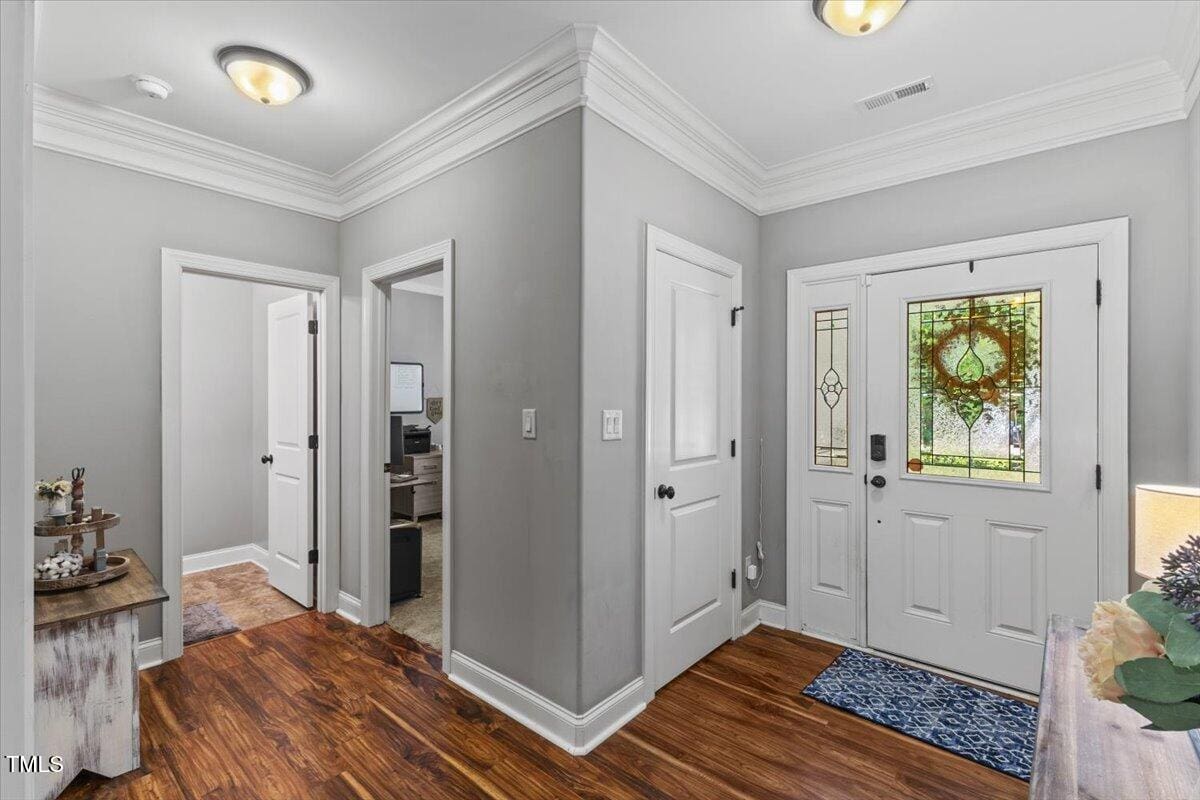 A hallway with wood floors, gray walls, crown molding, three white doors, and a front door with a decorative glass panel. There is a small table with decor and a blue rug at the entry.