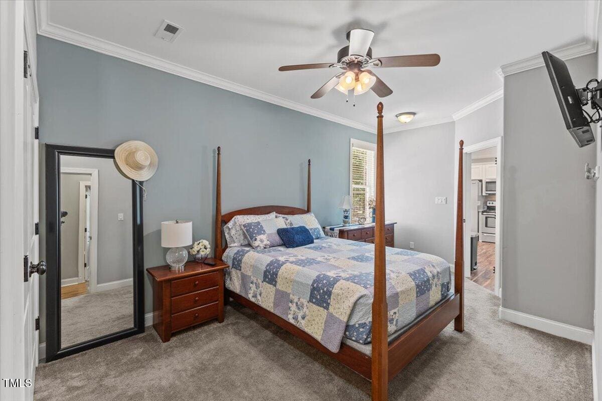 A bedroom with a wooden four-poster bed, patchwork quilt, two nightstands, a table lamp, a floor mirror with a hat, carpeted floor, ceiling fan with lights, and a wall-mounted TV. Kitchen visible through doorway.