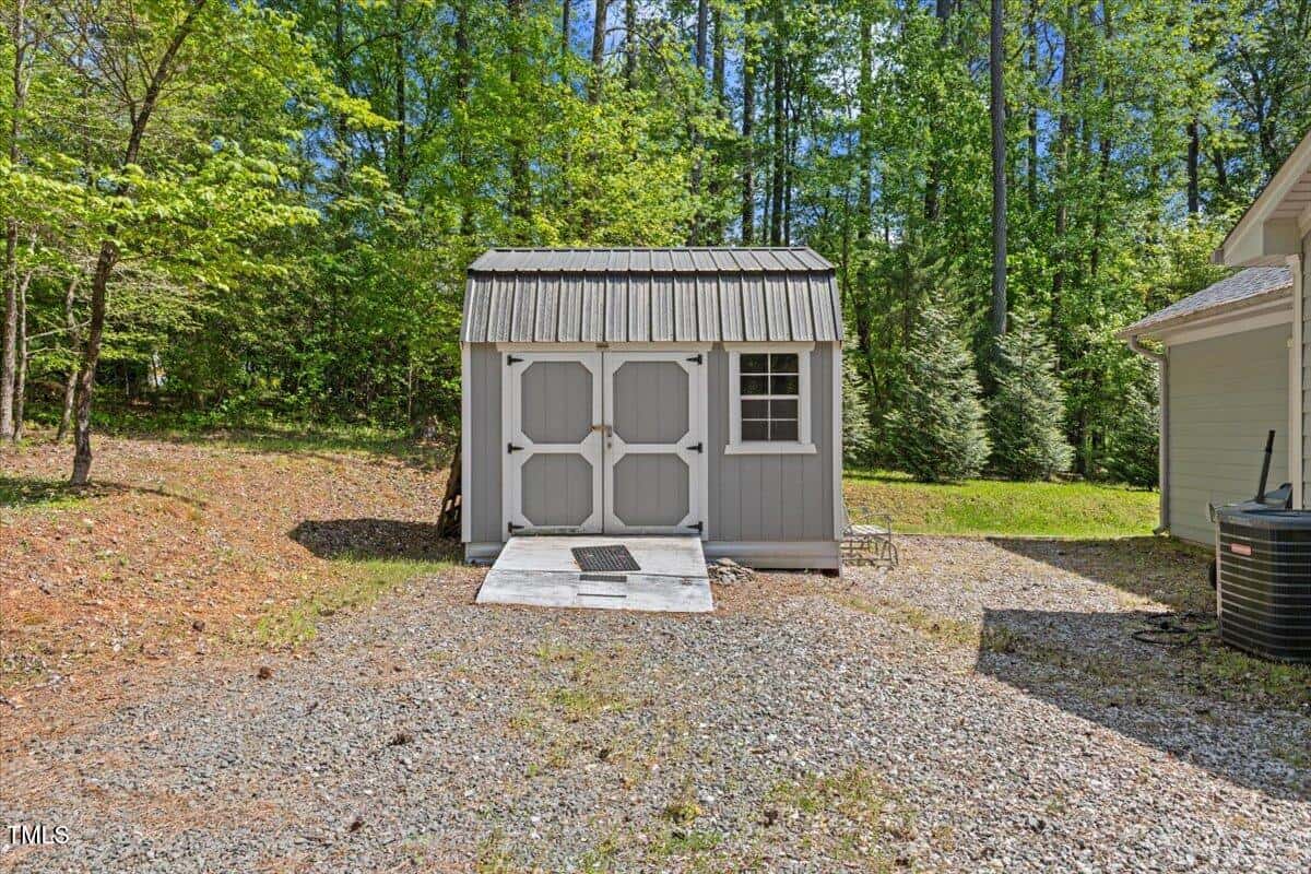 A small gray storage shed with double doors and a window sits on a concrete slab, surrounded by gravel and trees. A mat is in front of the entrance, and part of a house and an air conditioning unit are visible on the right.