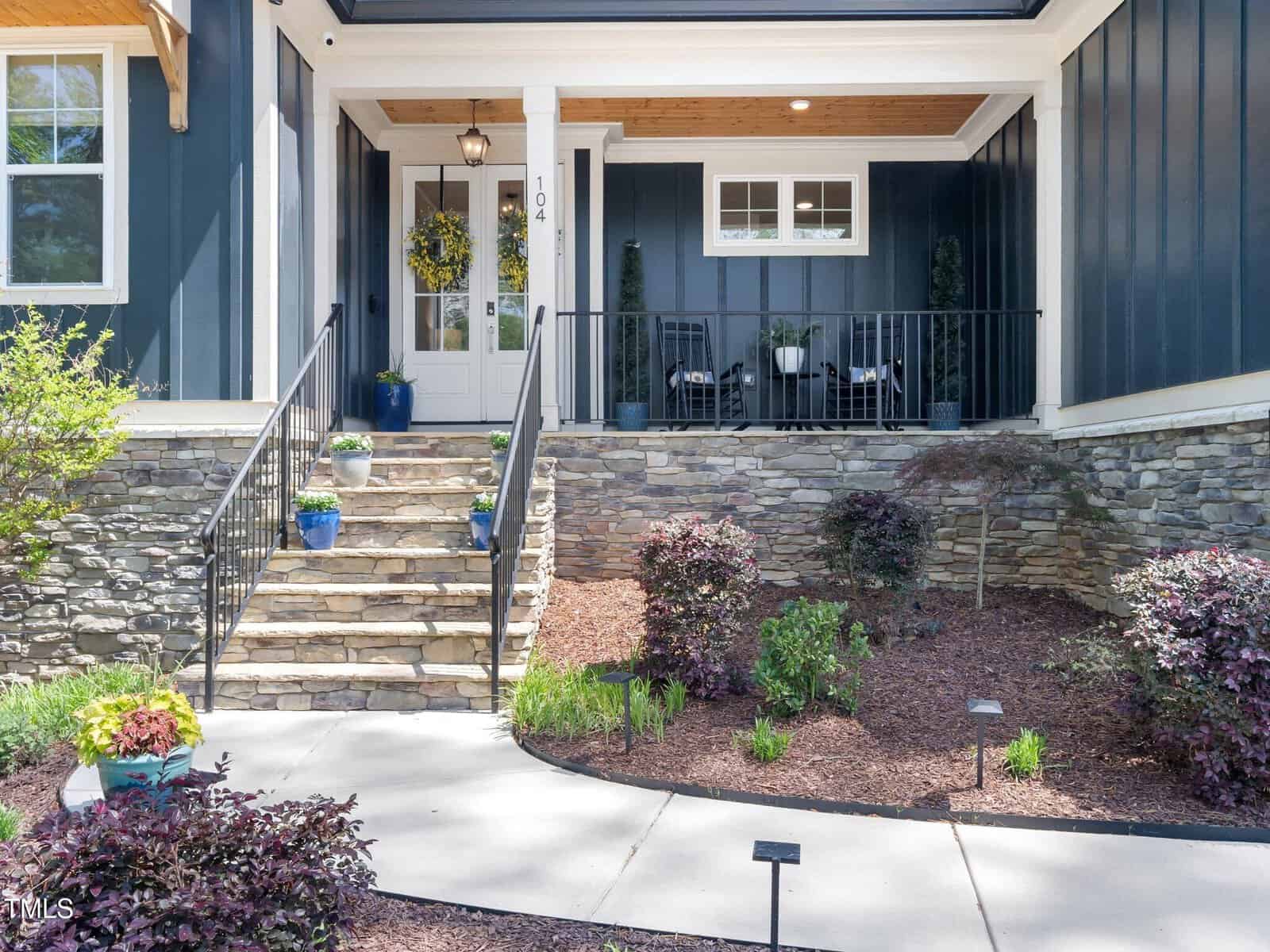 Front entrance of a house with stone steps, black railings, a white door with a wreath, potted plants, a porch with chairs, and landscaped garden beds with mulch and shrubs.