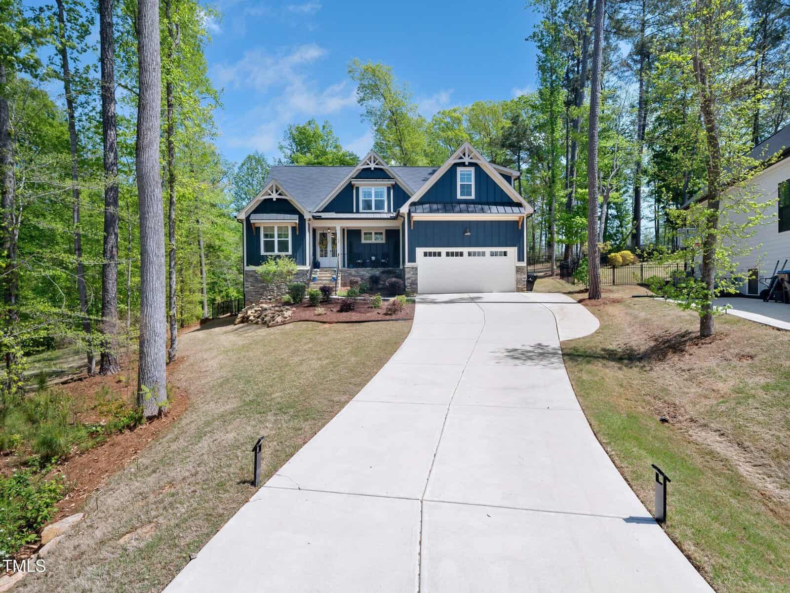 A modern two-story house with dark siding and a two-car garage sits at the end of a long, curved concrete driveway, surrounded by trees and landscaped with bushes on a sunny day.