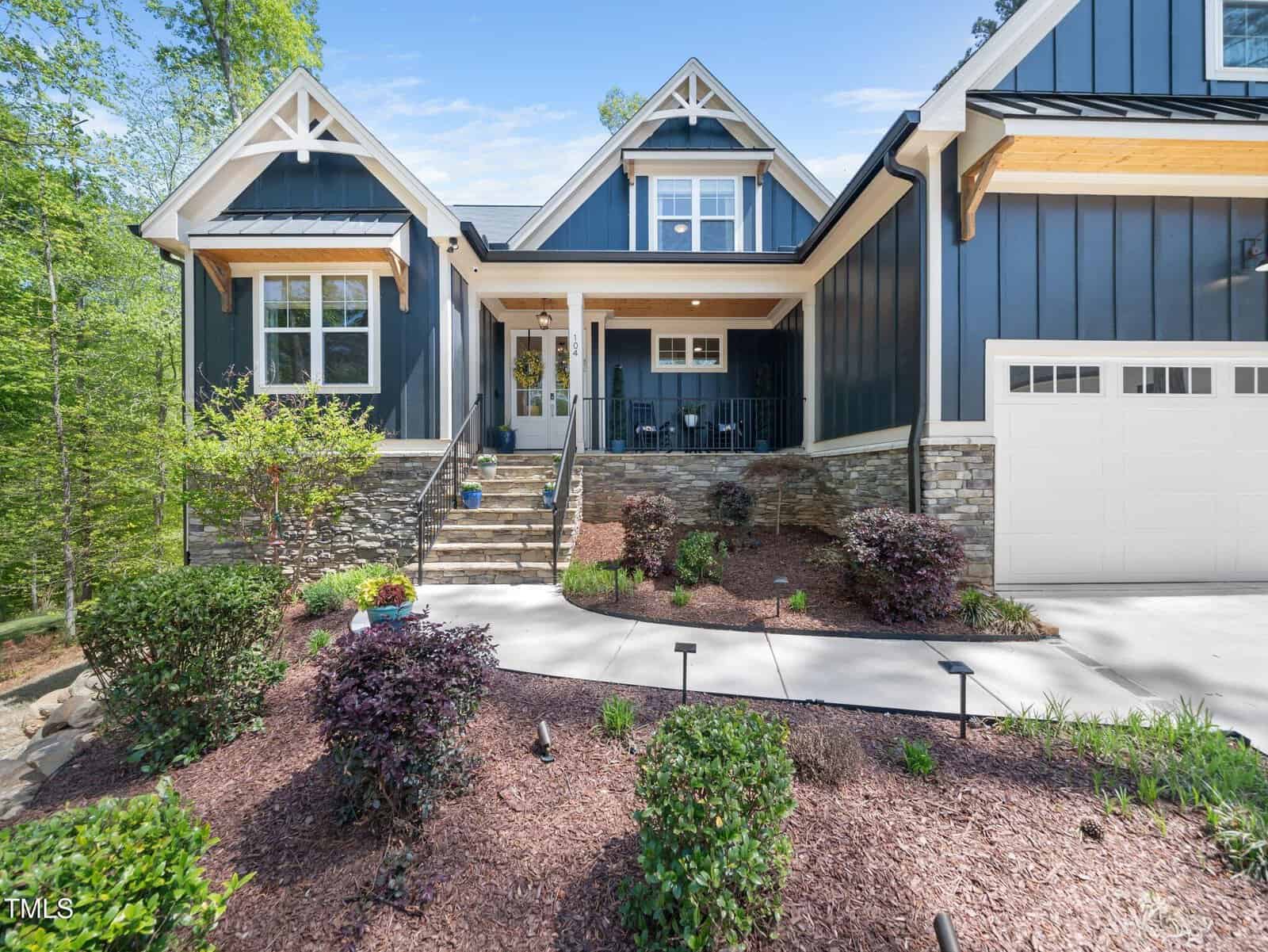 A modern blue house with white trim, stone accents, and a covered front porch. There are steps leading to the entrance, landscaped bushes, and a concrete walkway in the front yard under a clear sky.
