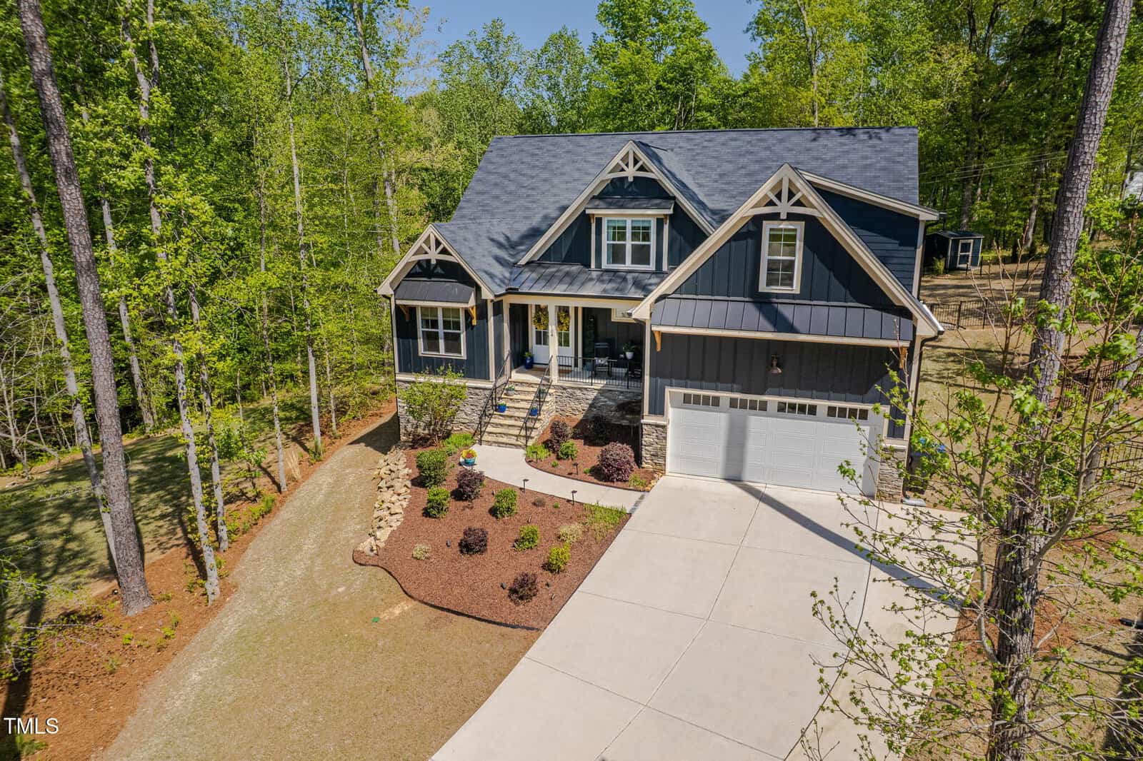 A two-story dark blue house with a covered front porch, dormer windows, and a two-car garage sits among tall trees. The front yard features landscaping with bushes, mulch, and a concrete driveway.