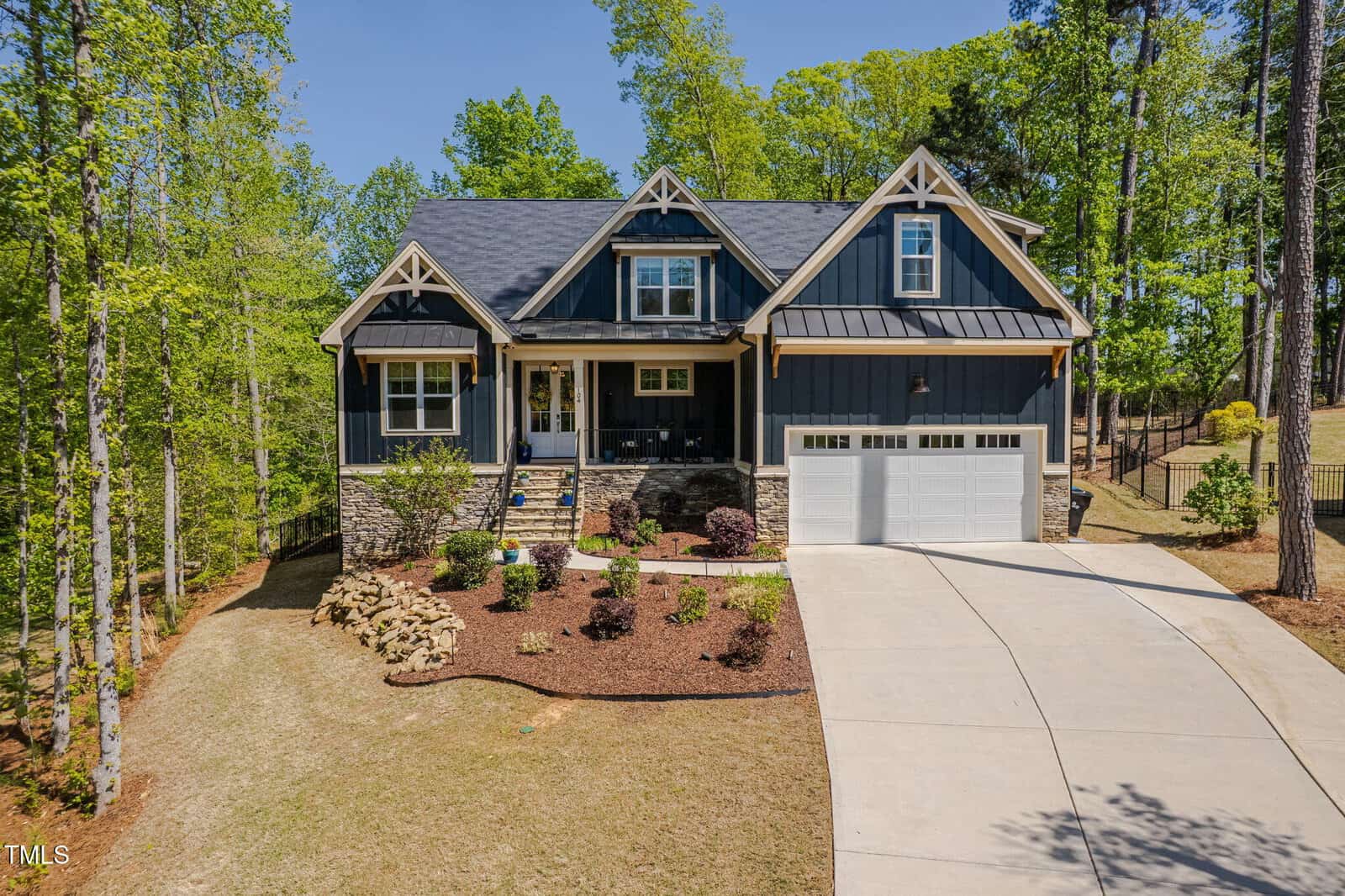 A two-story house with dark blue siding, white trim, a covered front porch, stone accents, and a two-car garage, surrounded by trees and landscaped yard with a curved concrete driveway.