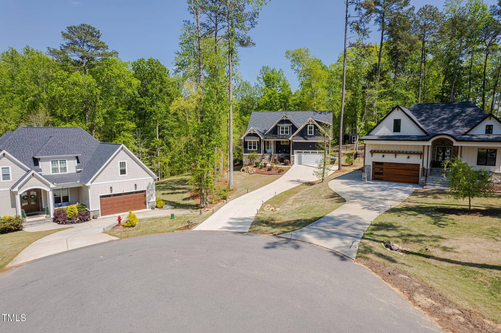 Three modern single-family homes with garages are situated on a cul-de-sac, surrounded by trees and greenery, each with paved driveways leading up from the street.