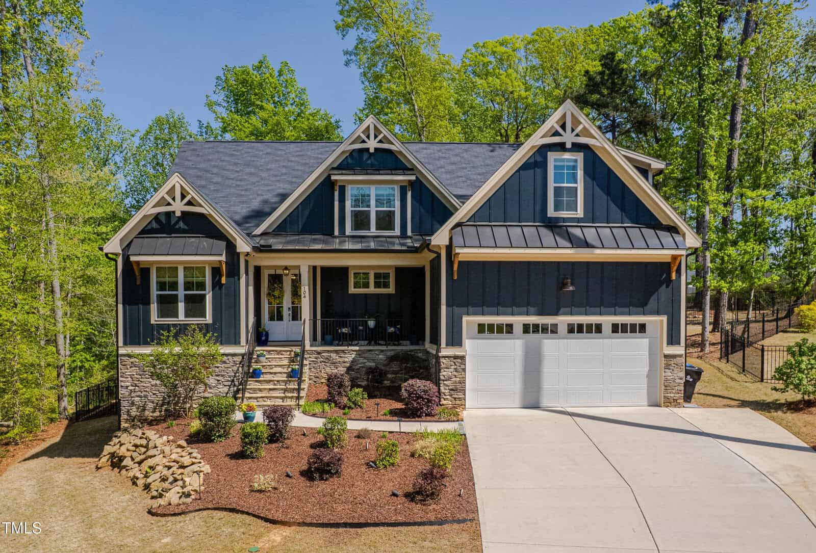 A two-story house with dark blue siding, white trim, a covered front porch, stone accents, and a double garage. The front yard is landscaped with shrubs, mulch, and a curved driveway. Tall trees surround the property.