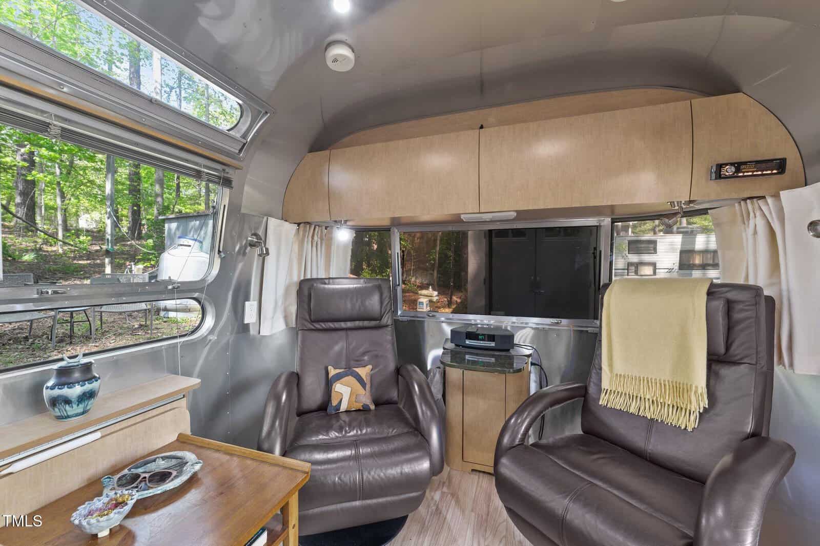 Two brown leather recliners face each other inside a modern Airstream trailer, with a window view of trees outside, light wood cabinetry, a table with a vase, and a yellow blanket draped over one chair.
