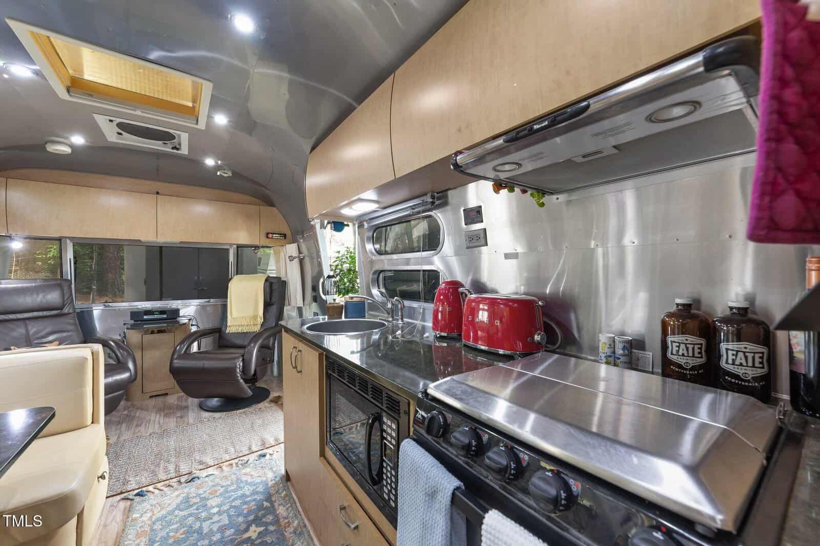 Modern RV kitchen with wood cabinets, stainless steel appliances, red toaster, red kettle, stove, sink, and lounge chairs in the background. Bright lighting and windows provide a spacious, cozy atmosphere.