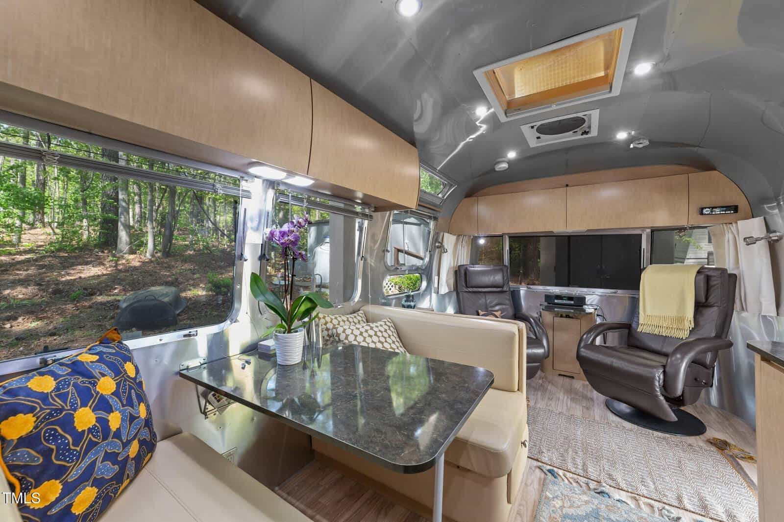 Interior of a modern Airstream trailer with a dining area, table, and cushioned seating, plus two recliner chairs. Large windows offer a forest view. There are light wood cabinets, a rug, an orchid, and throw pillows.