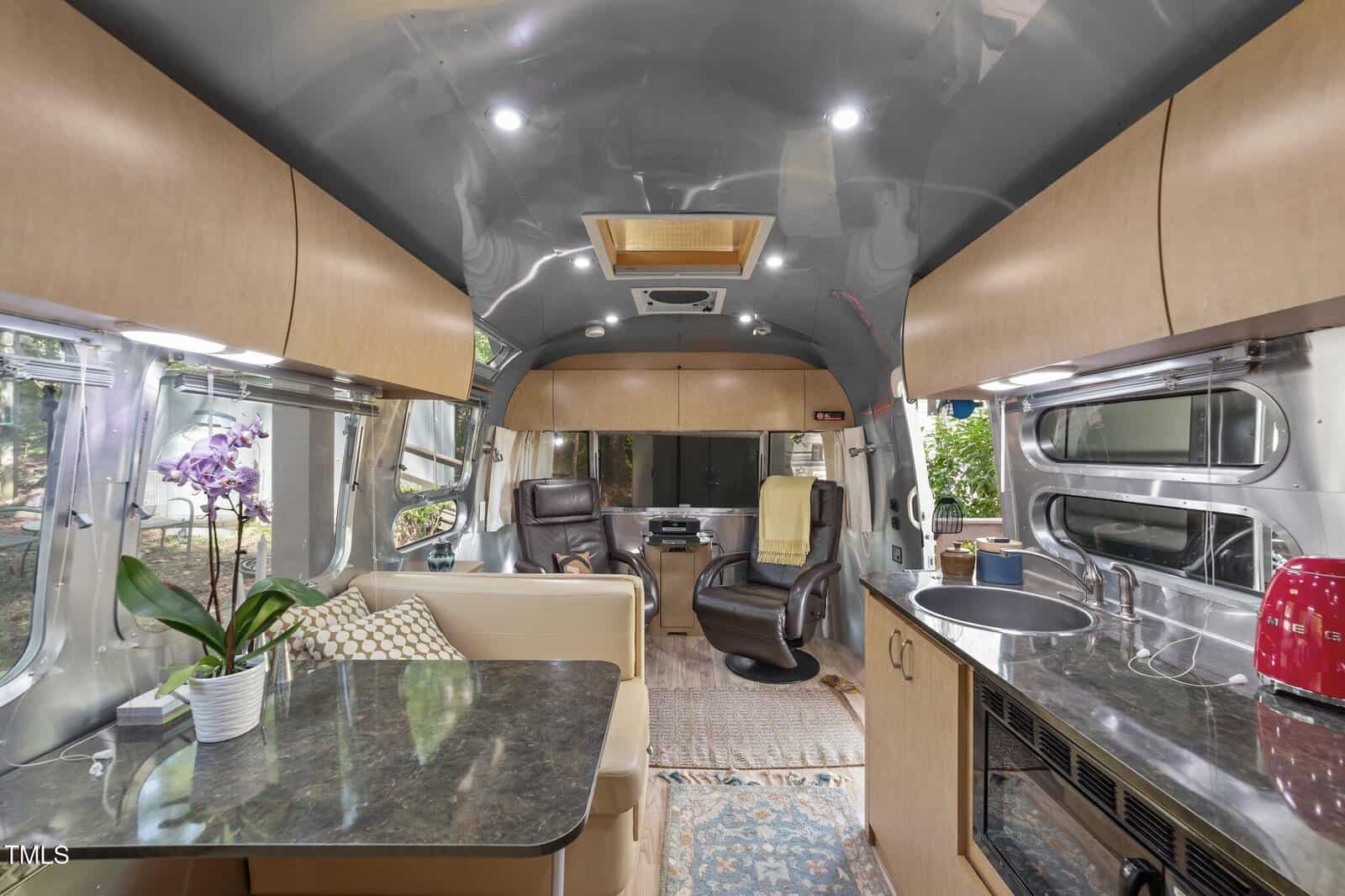 The interior of a modern RV with wood cabinets, a marble-look table holding an orchid, two leather chairs, a beige sofa, a kitchen area with a sink, stovetop, and red toaster, and large windows along the sides.