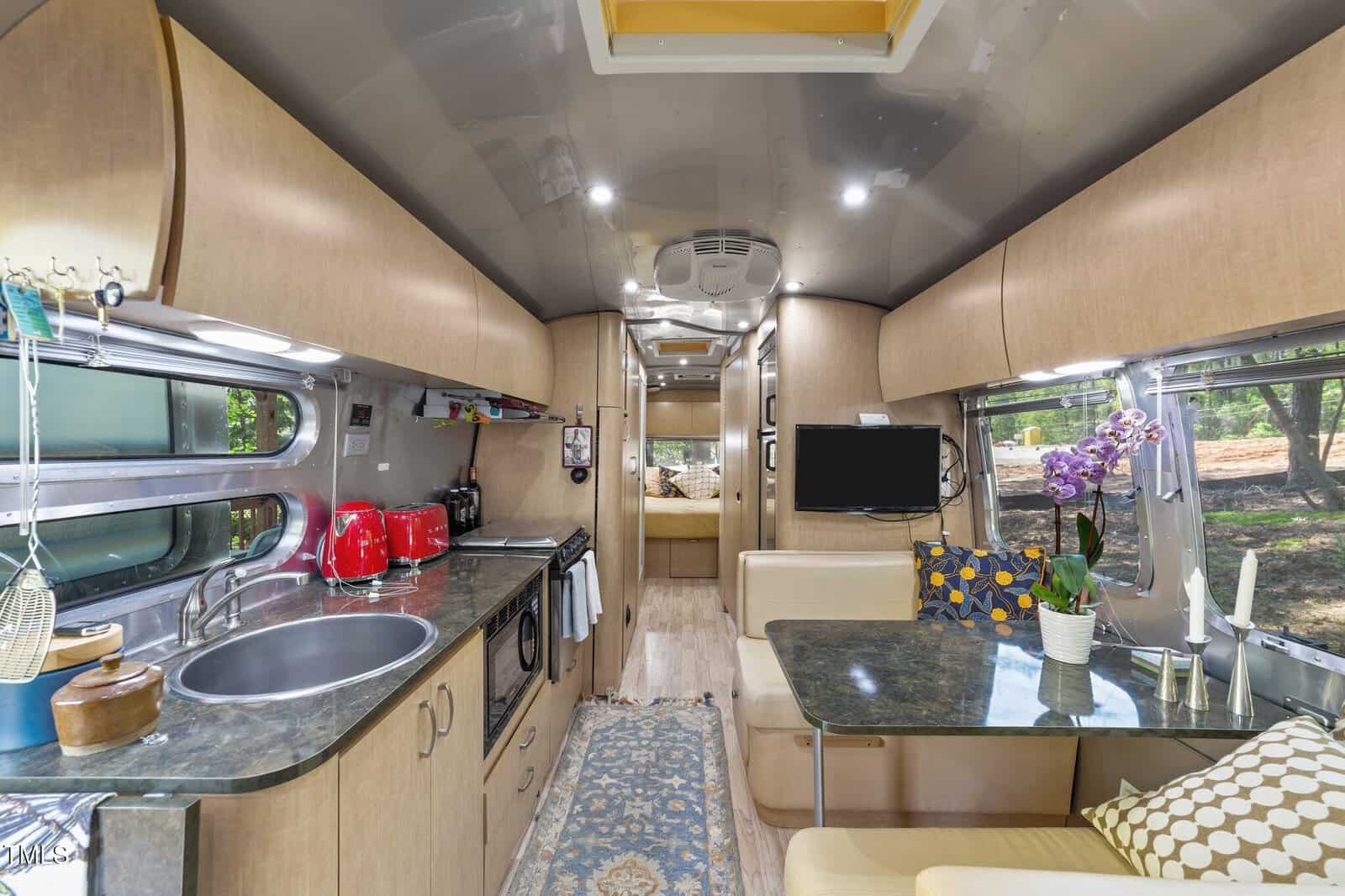 Interior of a modern RV featuring wood cabinetry, a small kitchen with sink and appliances, a dining area with table and cushions, a TV, and windows. The bedroom is visible in the background.