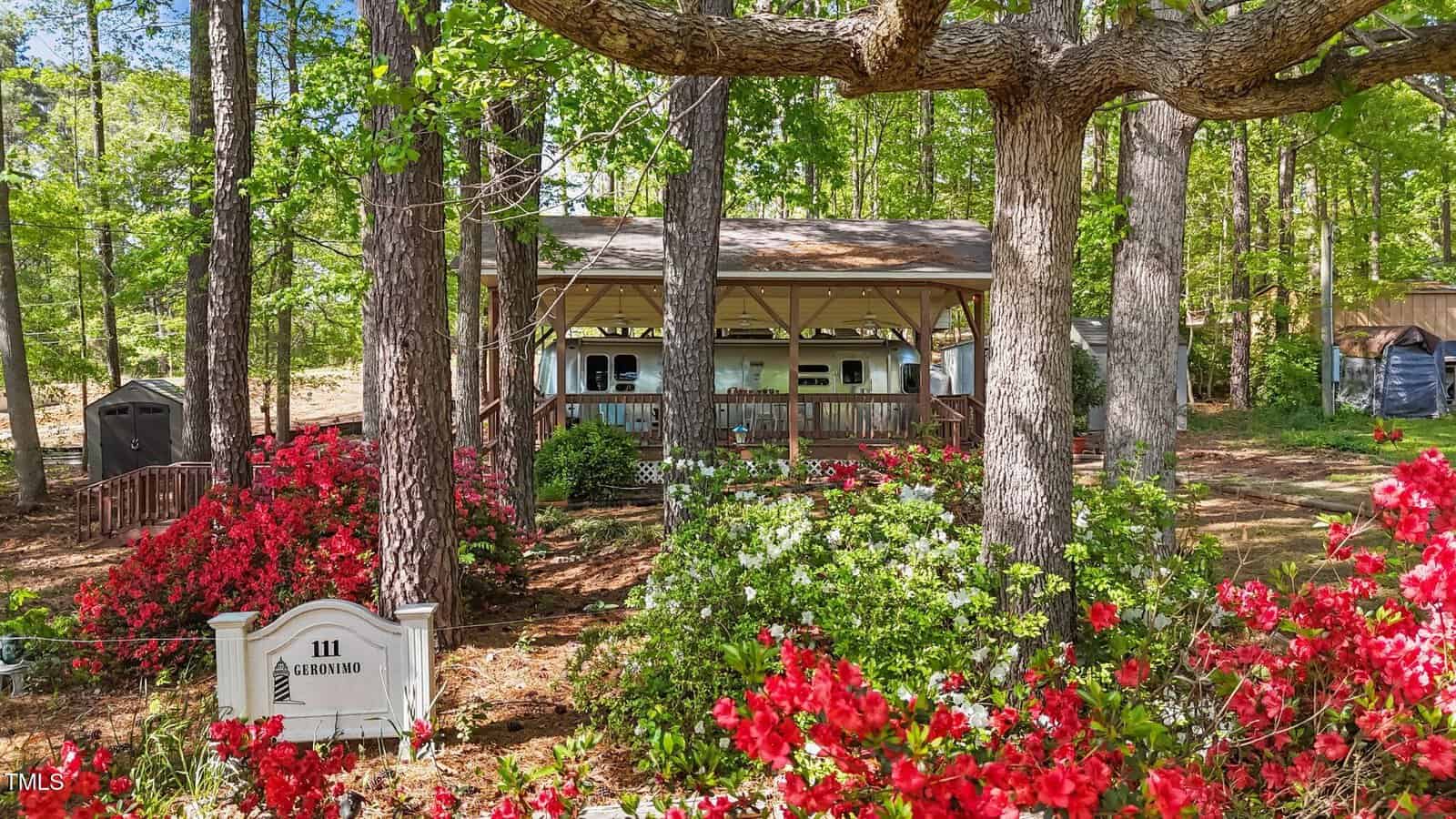 A cozy cabin with a covered porch sits among tall trees and vibrant red and white blooming flowers, with a sign that reads “111 GRINDSTONE” in the foreground.