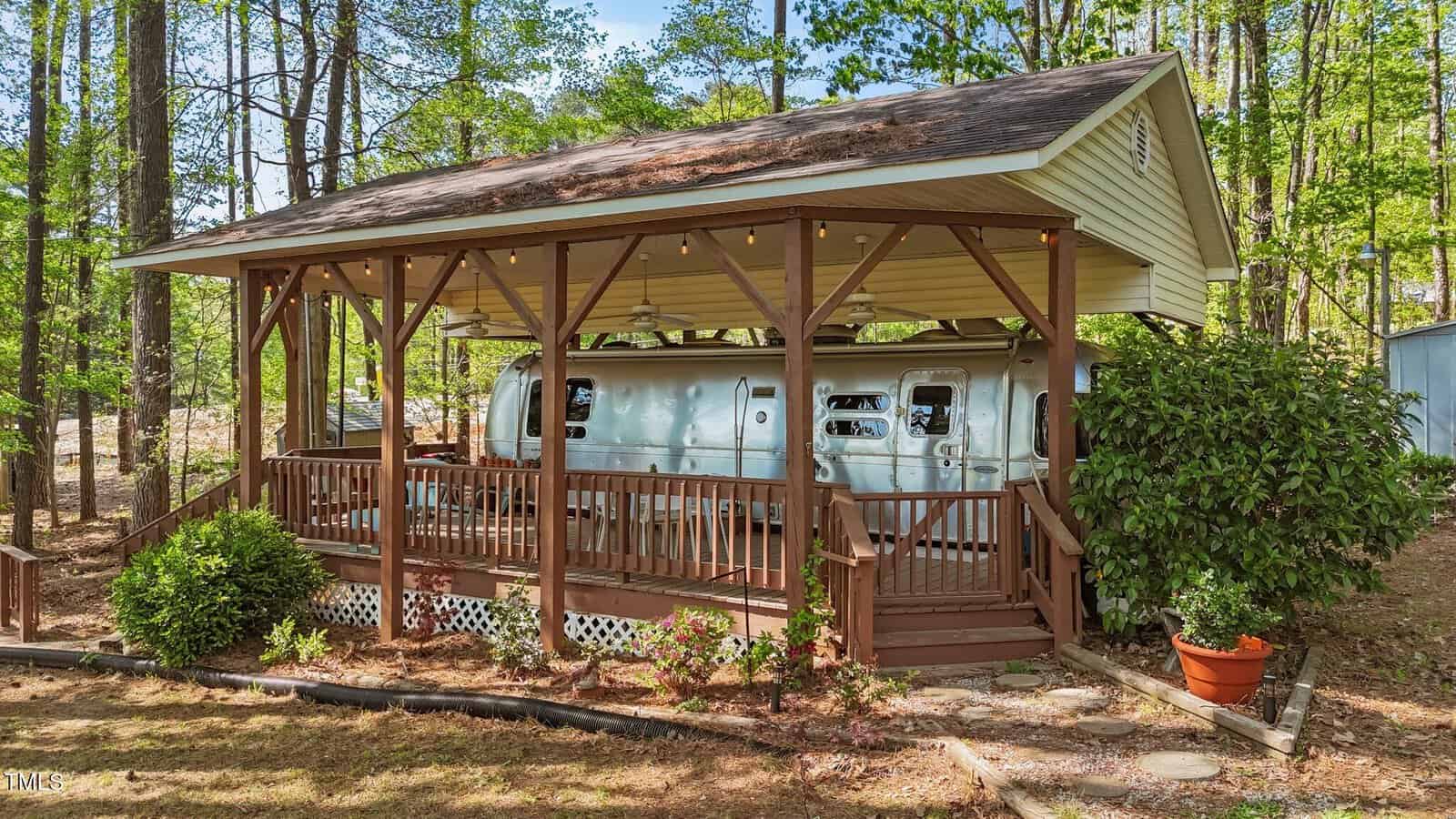 A silver Airstream trailer sits under a wooden pavilion with a pitched roof at 109-111 Geronimo Drive, Louisburg, surrounded by trees and greenery. A wooden deck with railings and stairs leads to the trailer, while potted and landscaped plants complete the inviting outdoor space.