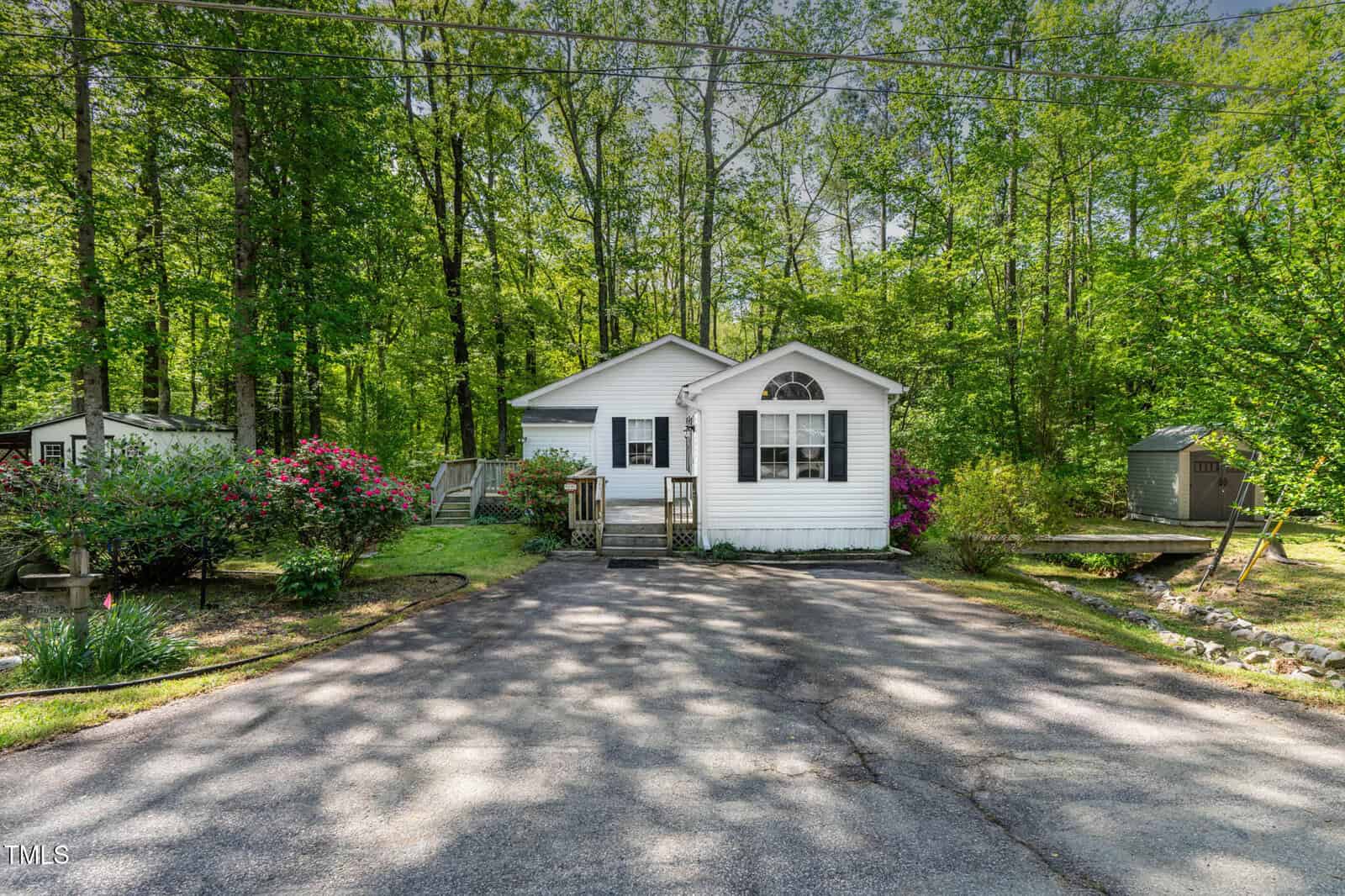 Small white house with a front porch, surrounded by lush green trees and gardens, with a paved driveway leading up to the entrance. Sheds are visible on both sides of the house.