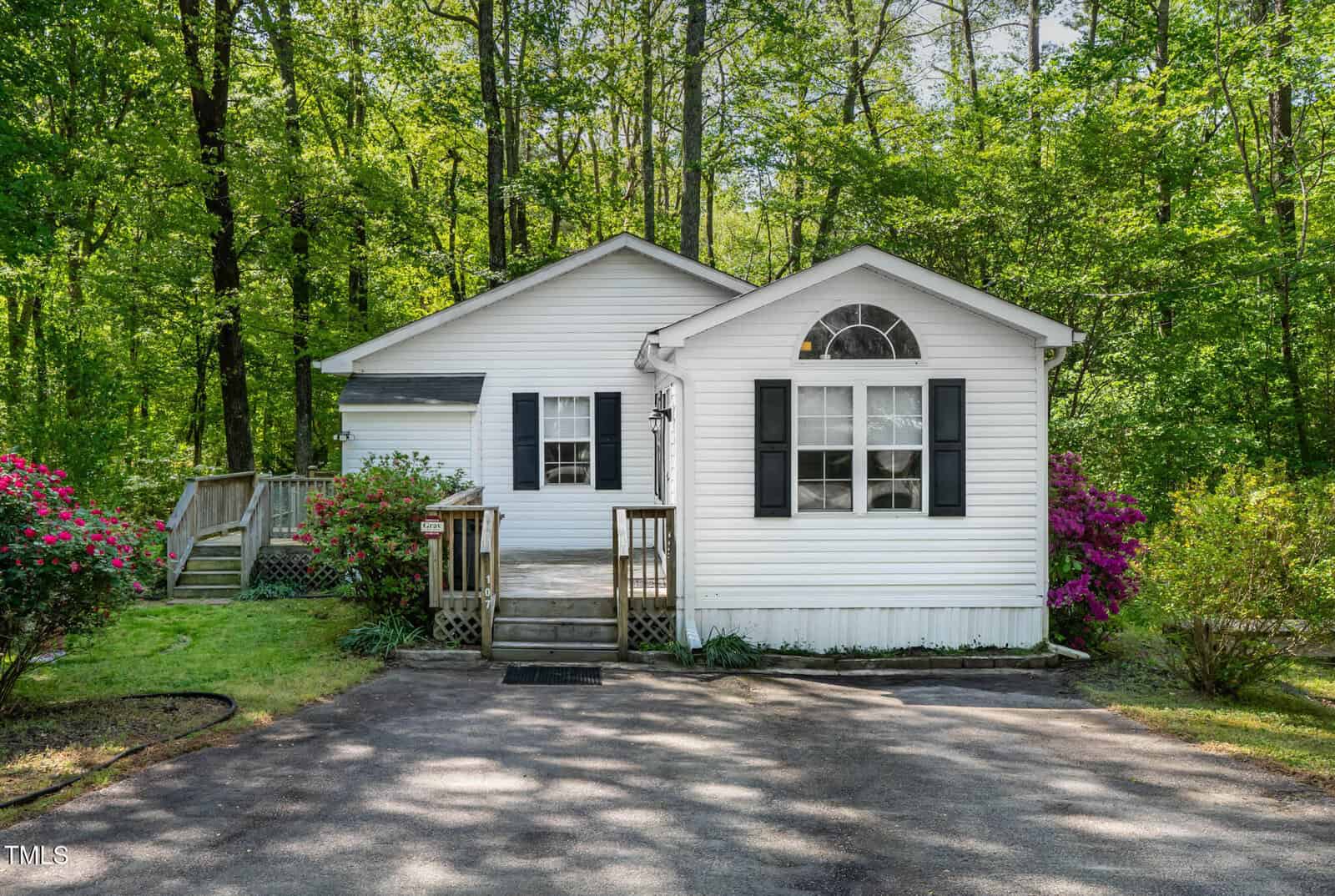 A small white house with black shutters and a front porch sits surrounded by green trees and blooming pink bushes, with a paved driveway in front.