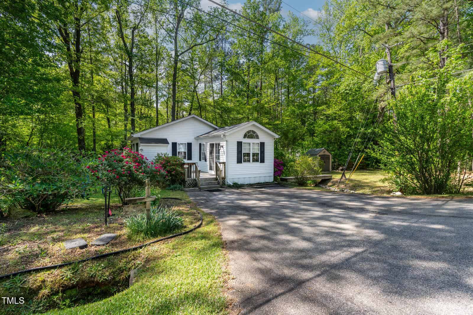 A small white house with a front porch sits surrounded by lush green trees and bushes. There is a paved driveway leading up to the house, and a garden with blooming flowers beside the driveway.