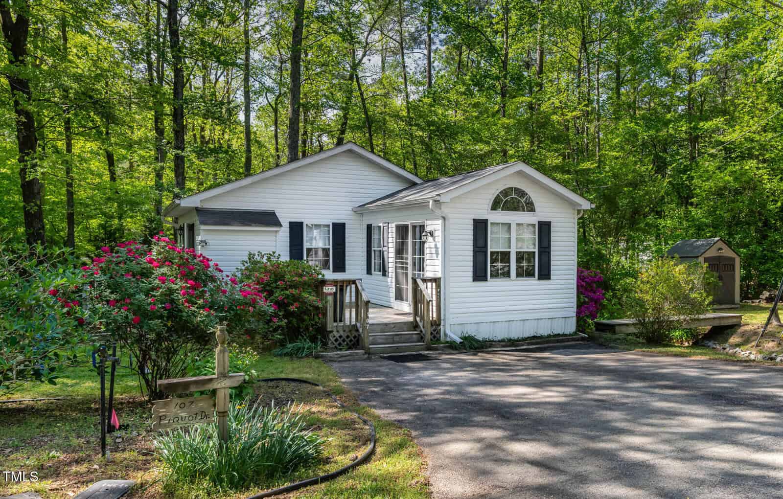 A small white house with dark shutters sits among lush green trees and flowering bushes, with a paved driveway and a small wooden shed in the background. Sunlight filters through the trees, creating a peaceful atmosphere.