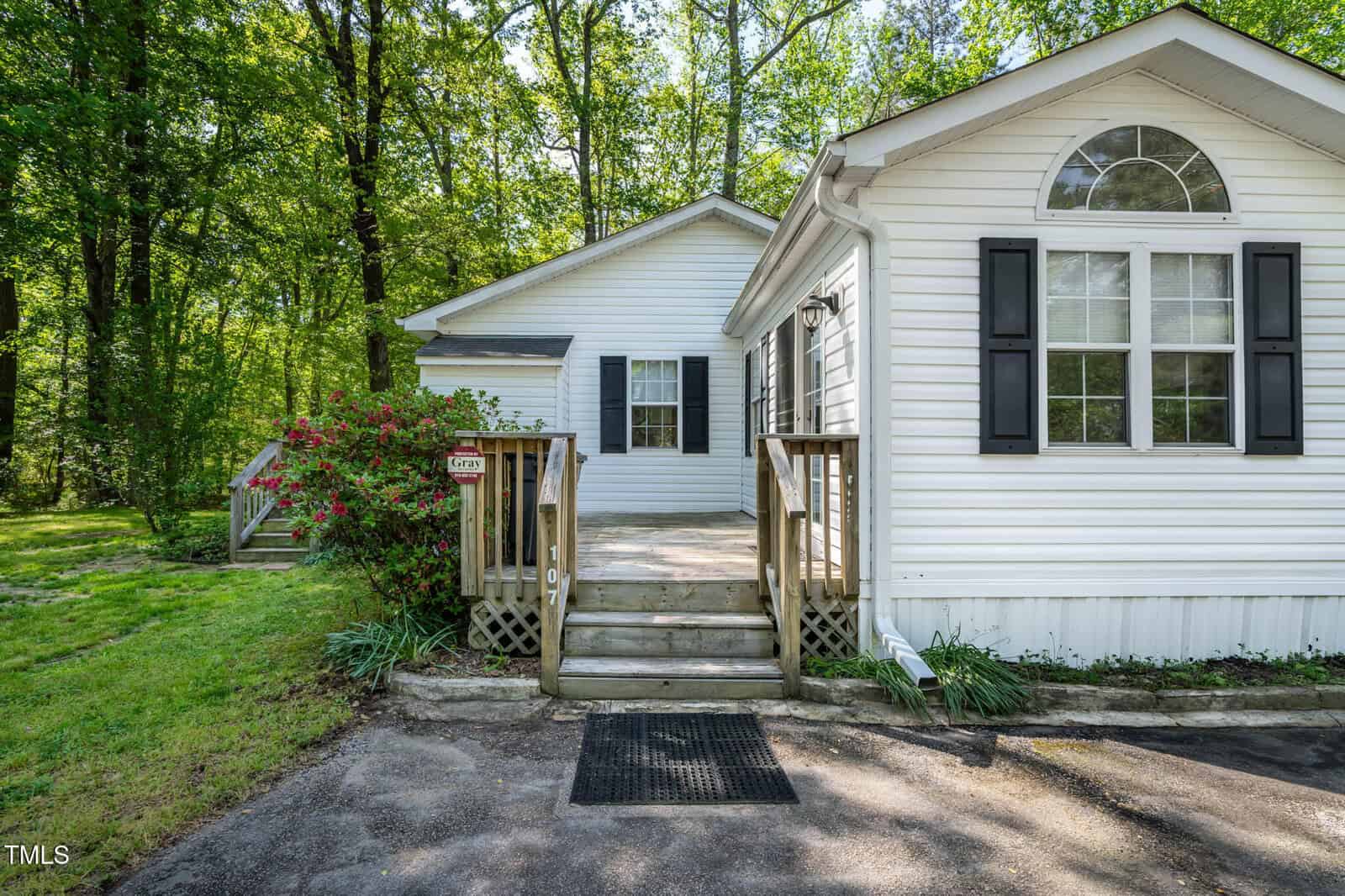 A white single-story house with black shutters and large windows, surrounded by trees and greenery. Wooden steps lead to the front door, with a small porch and driveway visible.