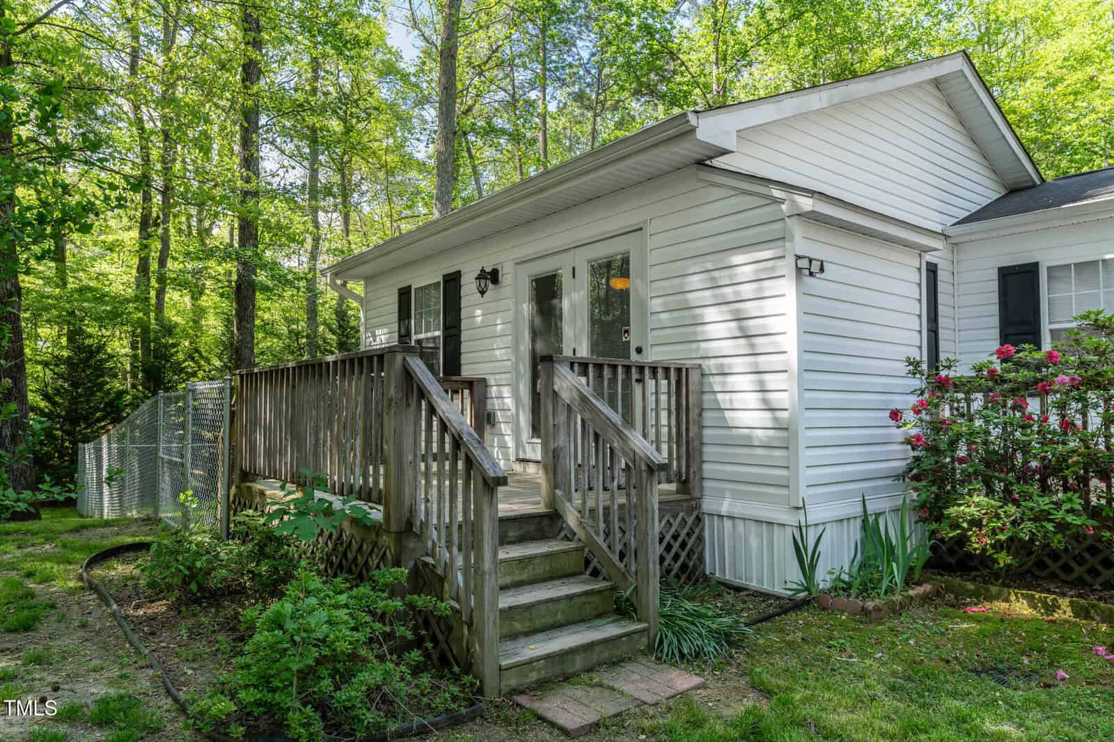 A small white house with horizontal siding, a wooden porch with stairs, and a railing. The yard is grassy with shrubs and flowers, and the house is surrounded by tall trees under bright sunlight.