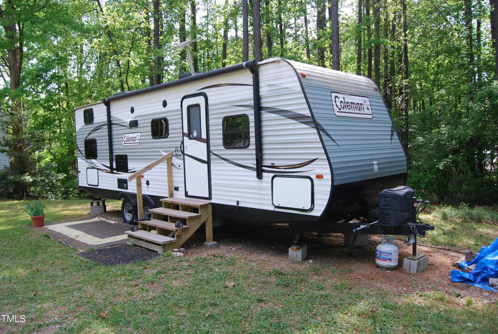 A white and gray Coleman travel trailer with steps leading to the door is parked in a wooded area. A propane tank and grill are in front, and a potted plant sits on a small patio to the side.
