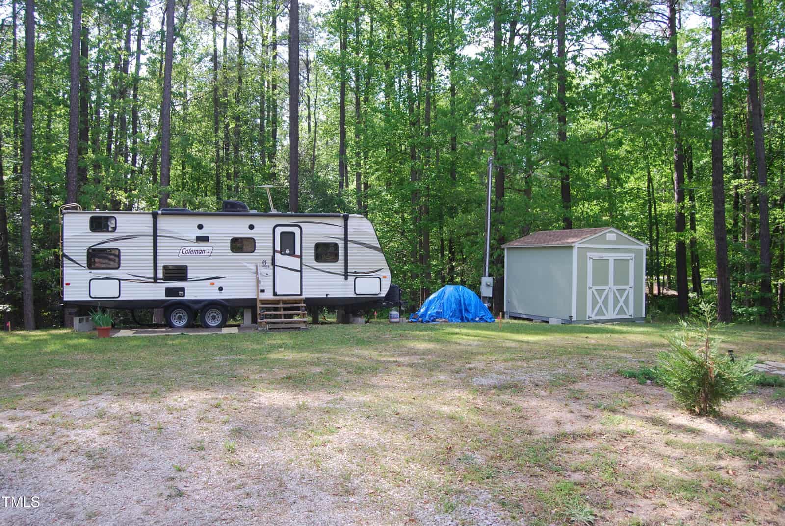 A white travel trailer is parked in a wooded area next to a small gray shed. A blue tarp covers items beside the shed, and tall green trees surround the open, grassy clearing.