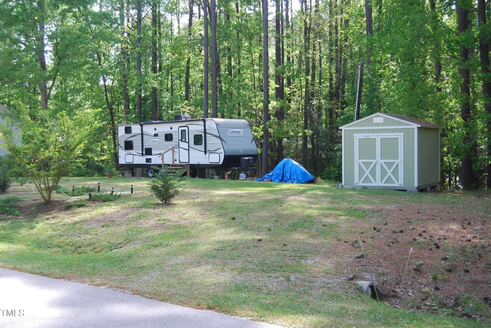 A travel trailer and a green storage shed sit on a grassy lot surrounded by tall trees. A blue tarp covers items near the shed, and the area is bordered by a road in the foreground.