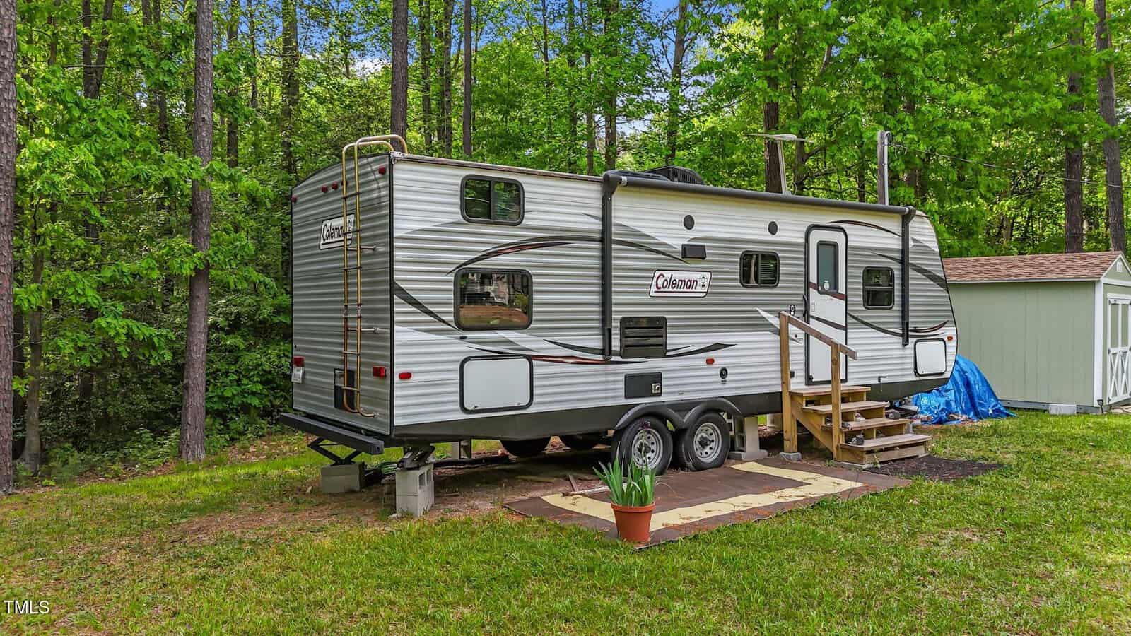 A gray Coleman travel trailer with steps and a small porch mat is parked on grass near a wooded area. A potted plant sits beside the trailer, and a small shed and blue tarp are visible in the background.