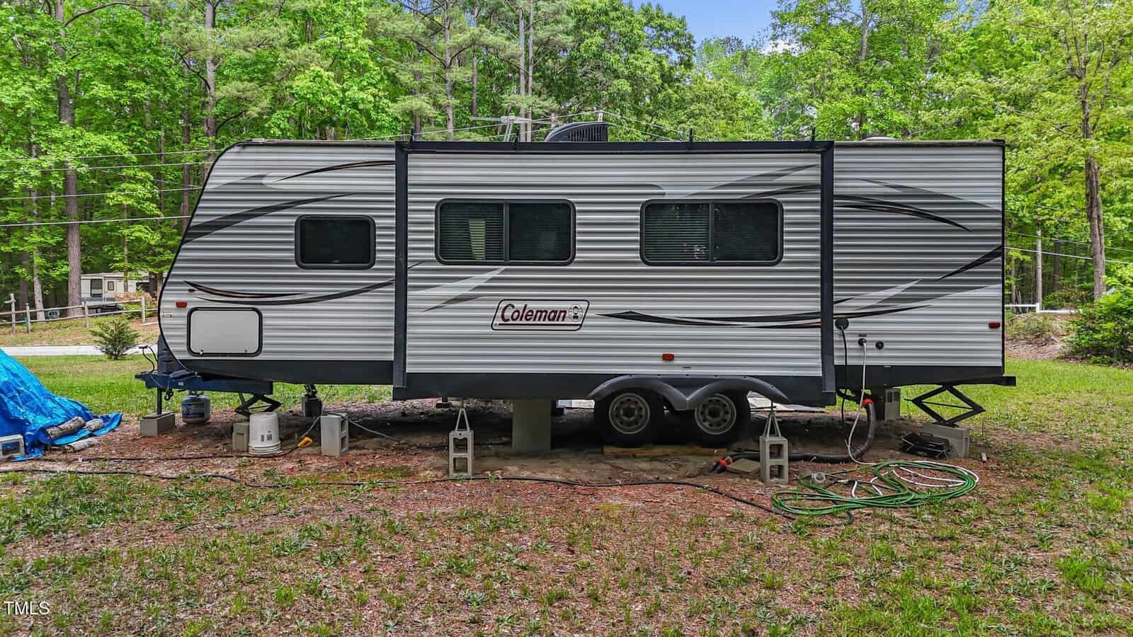 A silver Coleman travel trailer is parked on a grassy area, supported by cinder blocks. Trees and a few houses are visible in the background. Gardening tools and a hose are on the ground nearby.