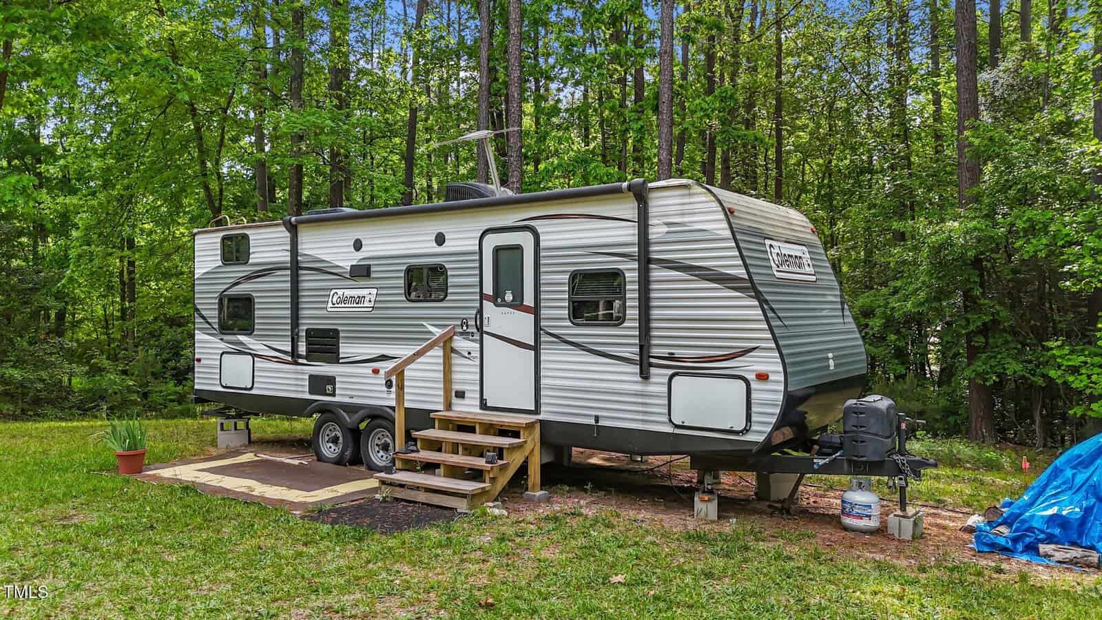 A silver Coleman travel trailer with two axles is parked on grass beside a wooded area. Wooden steps lead to the door, and a potted plant sits nearby. A tarp and two propane tanks are visible beside the camper.