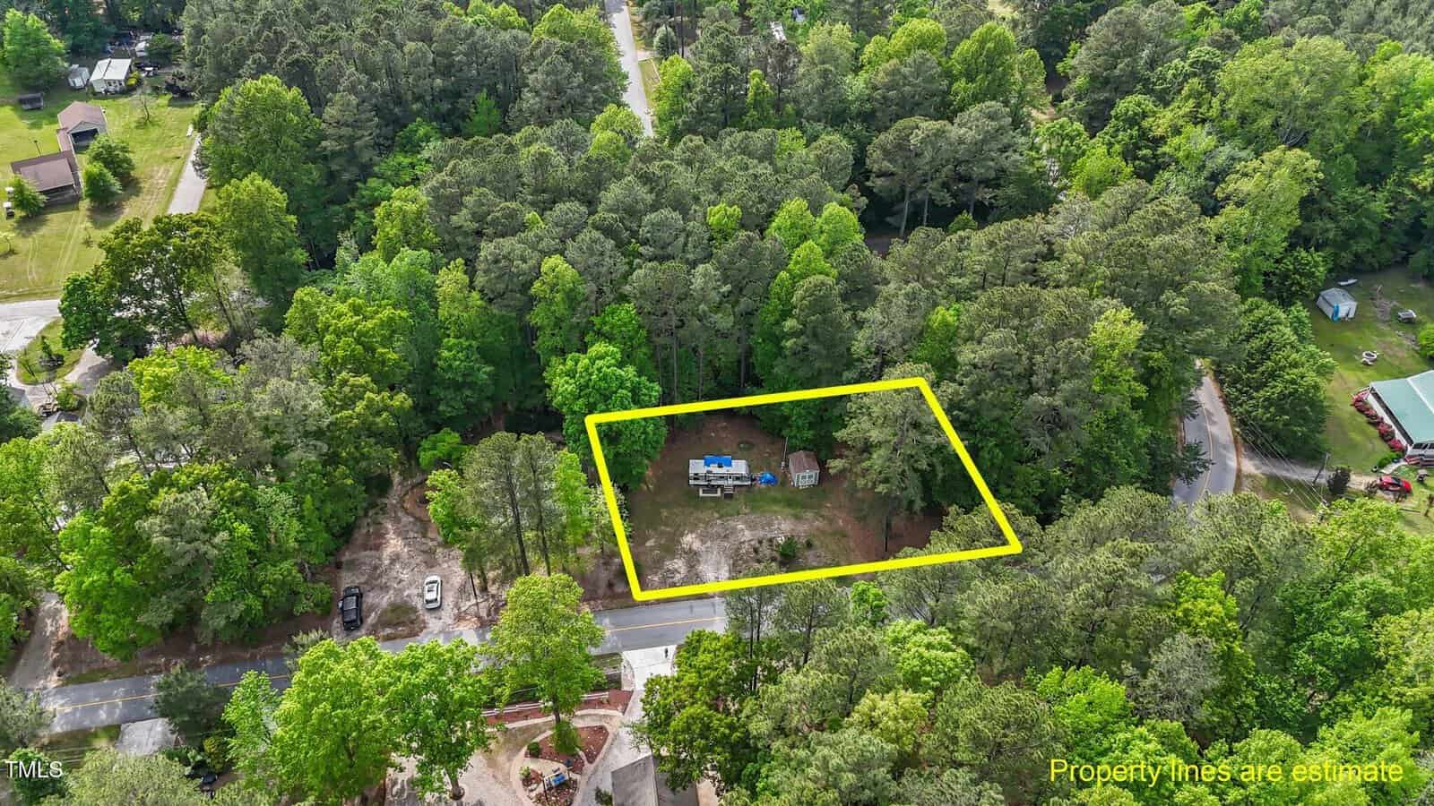 Aerial view of a wooded area with a rectangular plot outlined in yellow. Inside the outlined area are vehicles and some structures. The surrounding area is densely wooded, with roads and scattered houses visible.