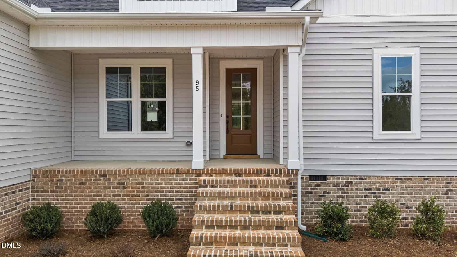 Front porch of a modern house with gray siding, a brown door with glass panels, brick steps, and small green shrubs planted along the brick foundation. The house number "95" is displayed beside the door.