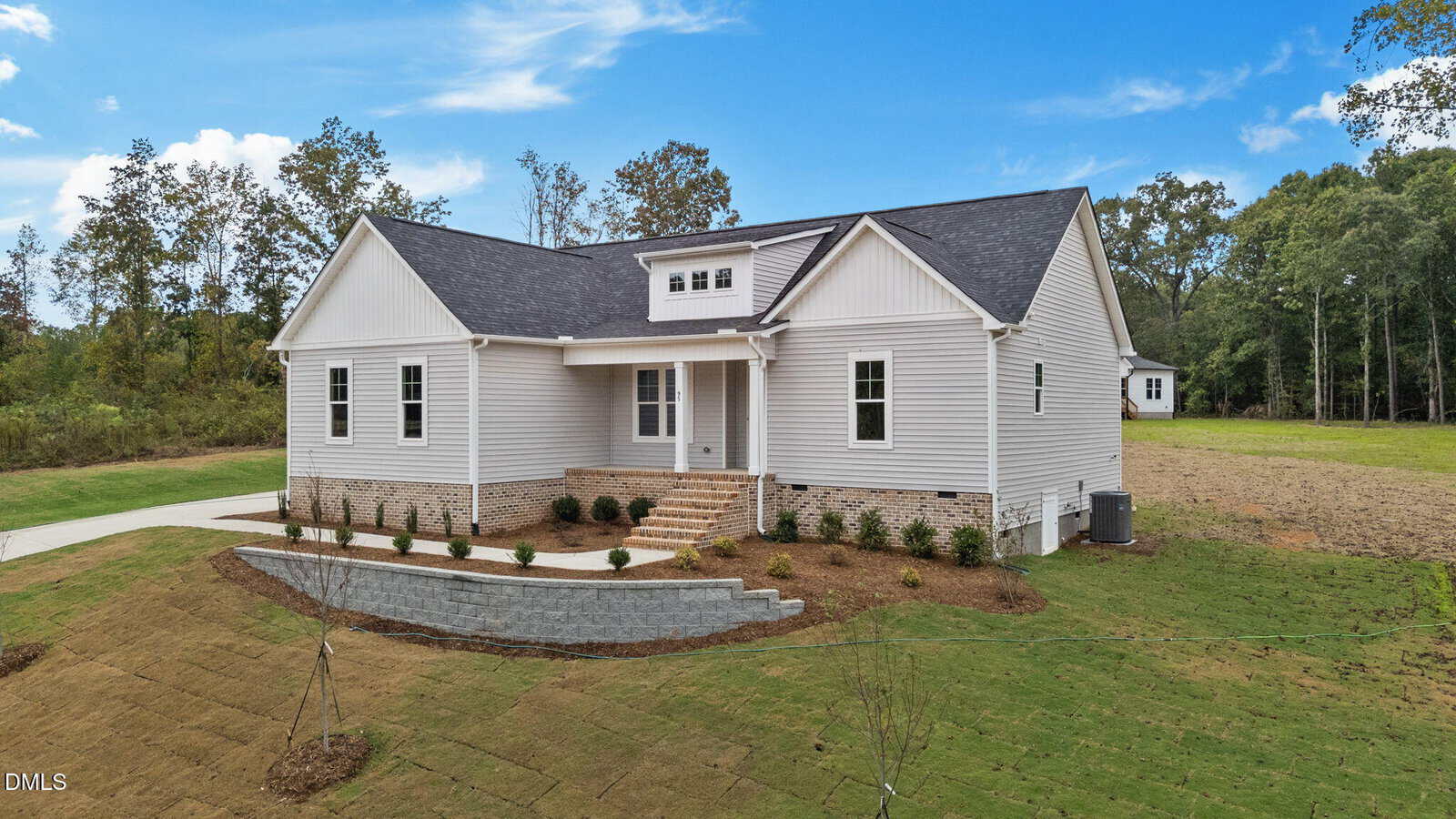 A new single-story white house with a covered front porch, brick steps, and a landscaped yard. The home sits on a large grassy lot bordered by trees under a bright blue sky with scattered clouds.