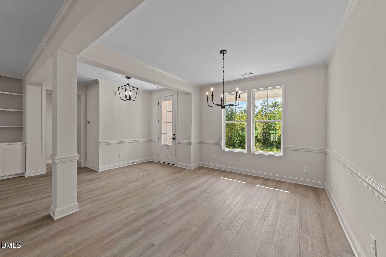 A bright, empty room with light wood floors, white walls, large windows, two modern black light fixtures, and built-in shelves, connected to another area by wide openings.