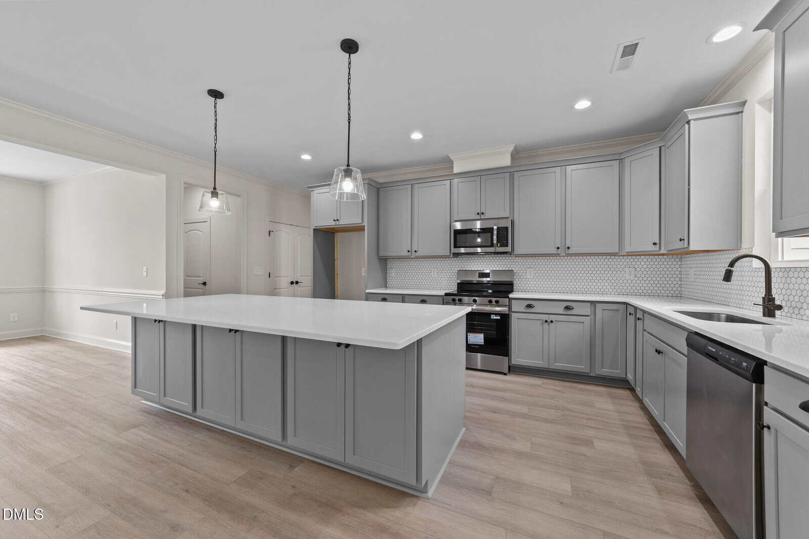 Modern kitchen with light gray cabinets, a large central island, stainless steel appliances, white countertops, tile backsplash, wood flooring, and pendant lighting. Bright, open layout with ample natural light.
