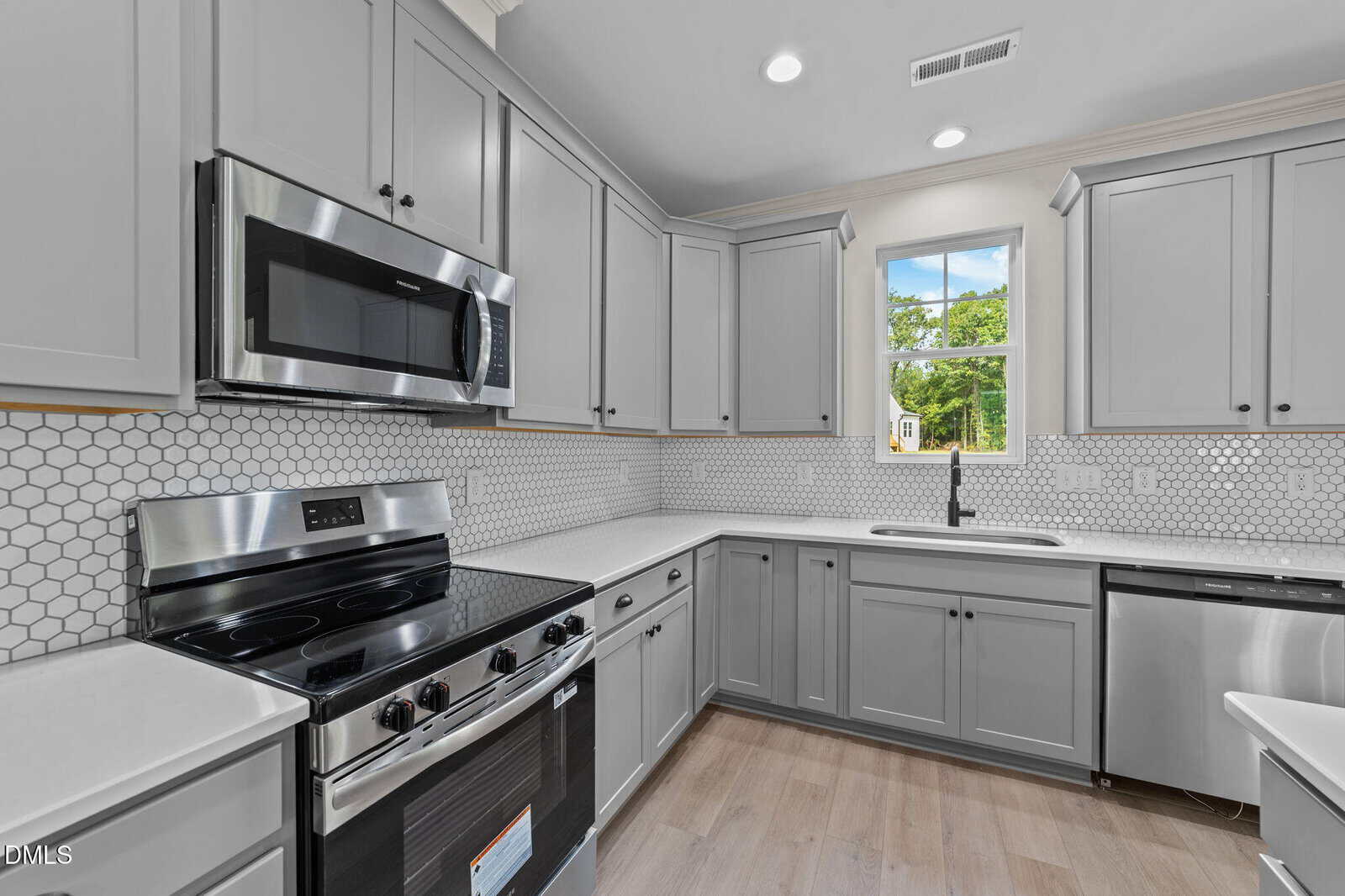 Modern kitchen with light gray cabinets, stainless steel appliances, a tile backsplash with a hexagonal pattern, a single window above the sink, and light wood flooring.