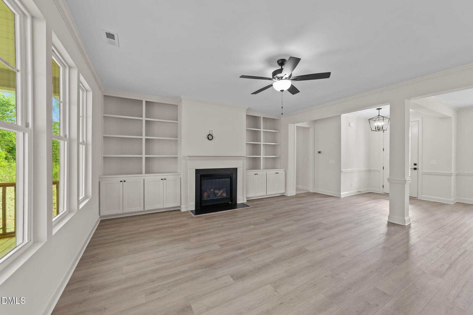 Bright, empty living room with light wood floors, built-in shelves, fireplace, ceiling fan, and windows. Open layout leads to entryway and dining area. White walls and trim throughout.