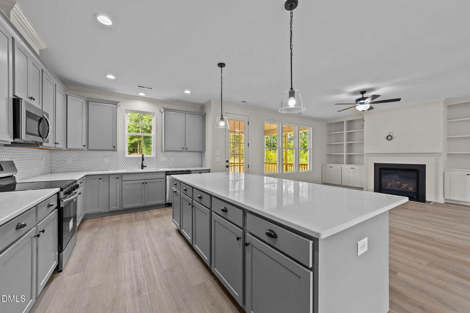Modern kitchen with gray cabinets, large white island, stainless steel appliances, pendant lights, light wood floors, and open living area featuring built-in shelves, fireplace, and ceiling fan. Large windows show a deck outside.