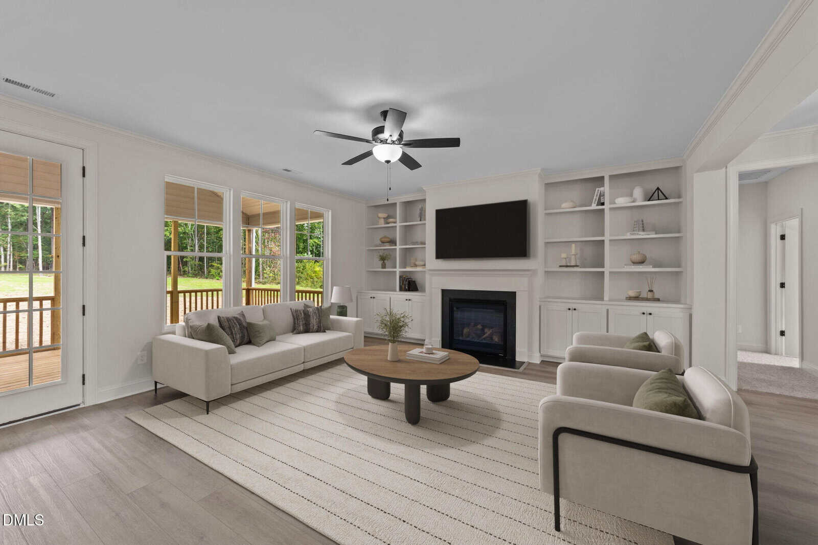 Modern living room with light sofas, a round wooden coffee table, built-in shelves, a fireplace, TV above the mantel, large windows, and glass doors opening to a porch with a view of green trees outdoors.