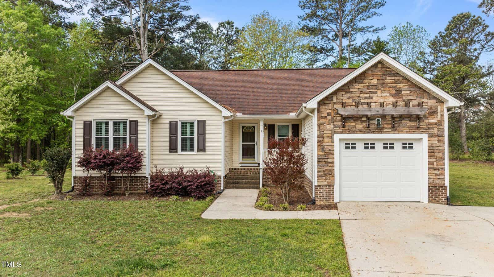 A single-story house with tan siding and stone accents, brown shingle roof, a front porch with steps, attached single-car garage, and a concrete driveway. There are small shrubs in front and trees in the background.