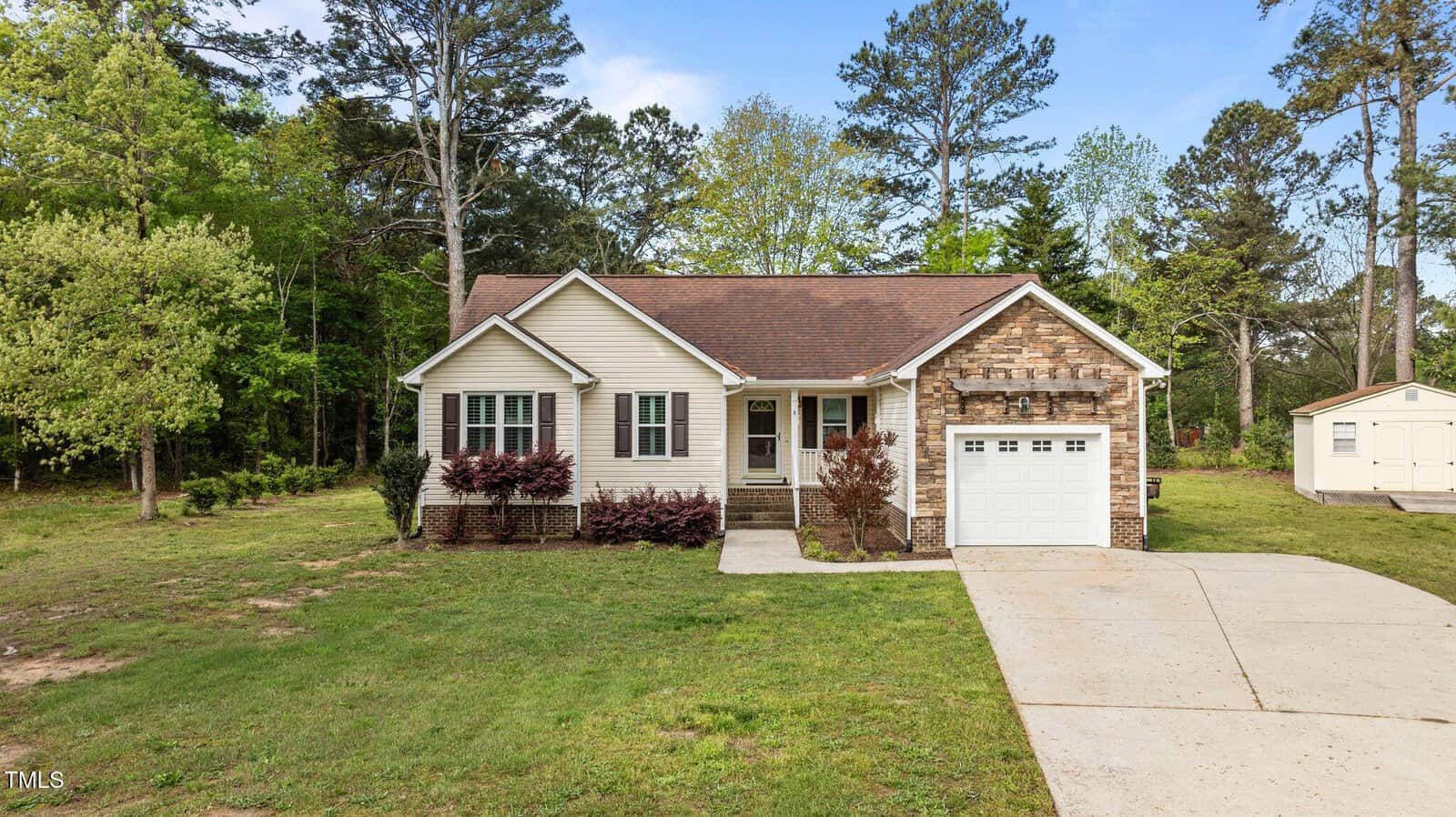 Single-story house with light-colored siding, brown shingle roof, and stone accents. Attached single-car garage, concrete driveway, small front porch, and minimal landscaping. Surrounded by trees and grass.