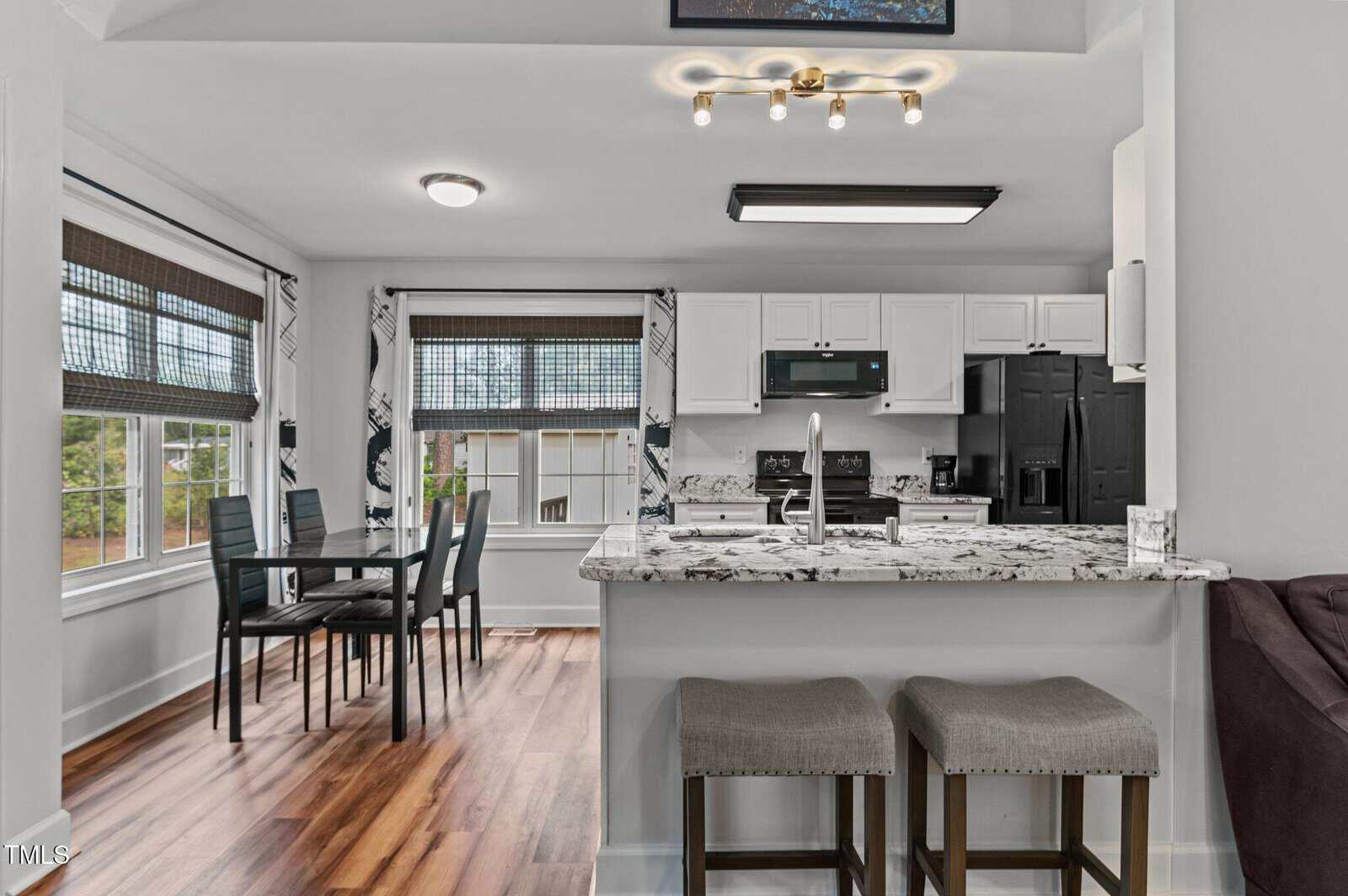 A modern kitchen with white cabinets, black appliances, and granite countertops, featuring a breakfast bar with two stools. Adjacent is a dining area with a glass table and four black chairs near large windows.