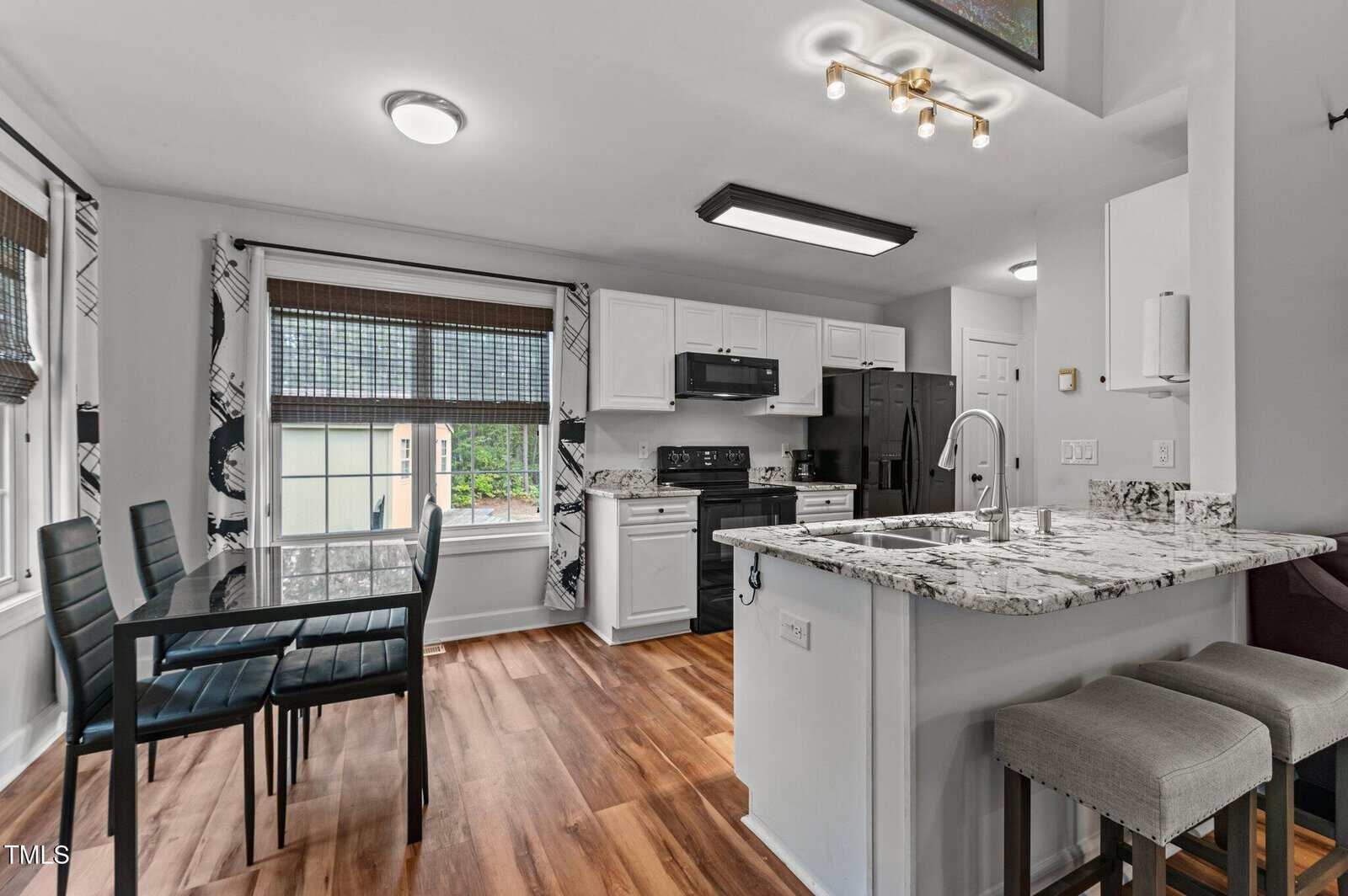 Modern kitchen and dining area with white cabinets, granite countertops, black appliances, a small dining table with four chairs, two bar stools at the counter, and wood flooring. Large windows provide natural light.