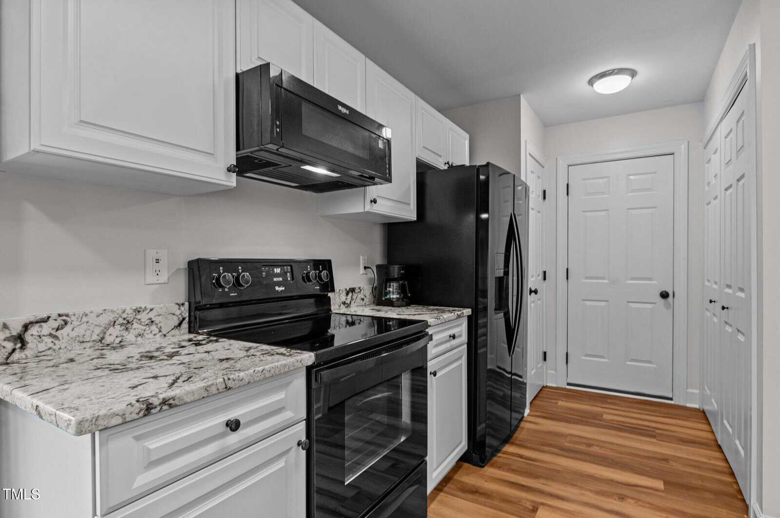 A modern kitchen features white cabinets, a black stove and microwave, a black refrigerator, granite countertops, and wood flooring. Multiple doors are visible in the background under ceiling light.